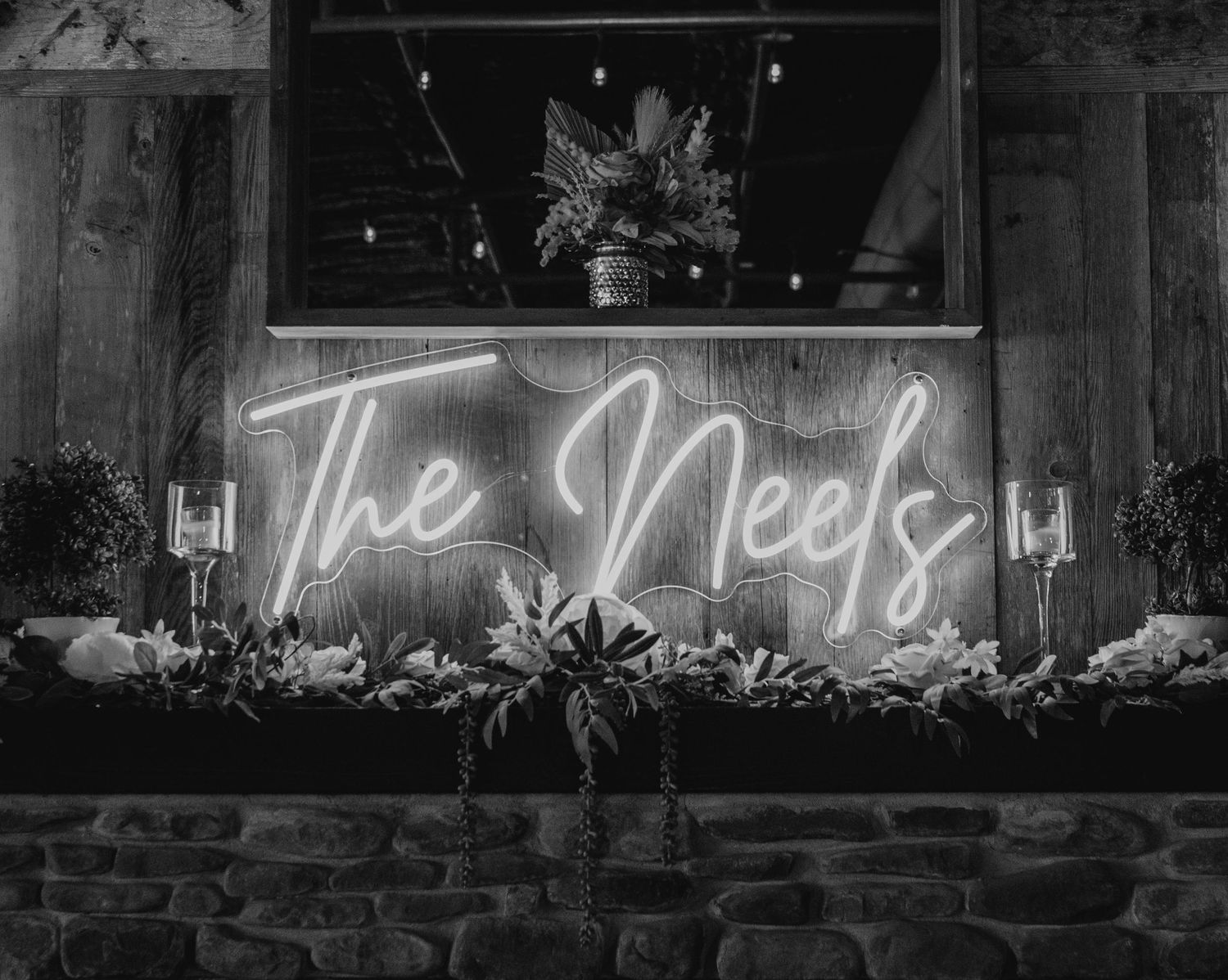 A glowing neon sign reading 'The Neels' mounted on a brick wall with decorative plants in black and white.