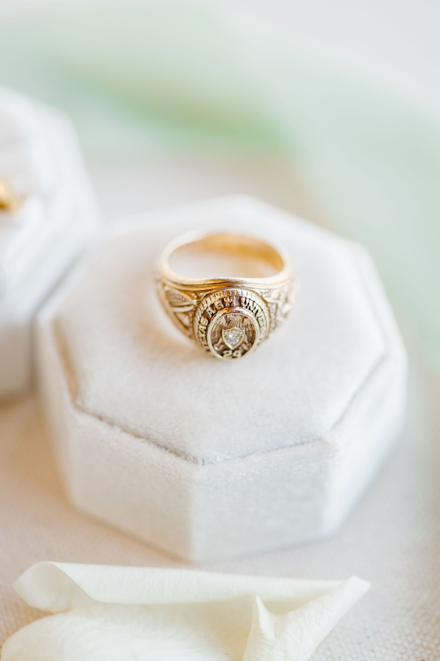 Vintage-style gold class ring with ornate details sits on white hexagonal box against soft white fabric backdrop.