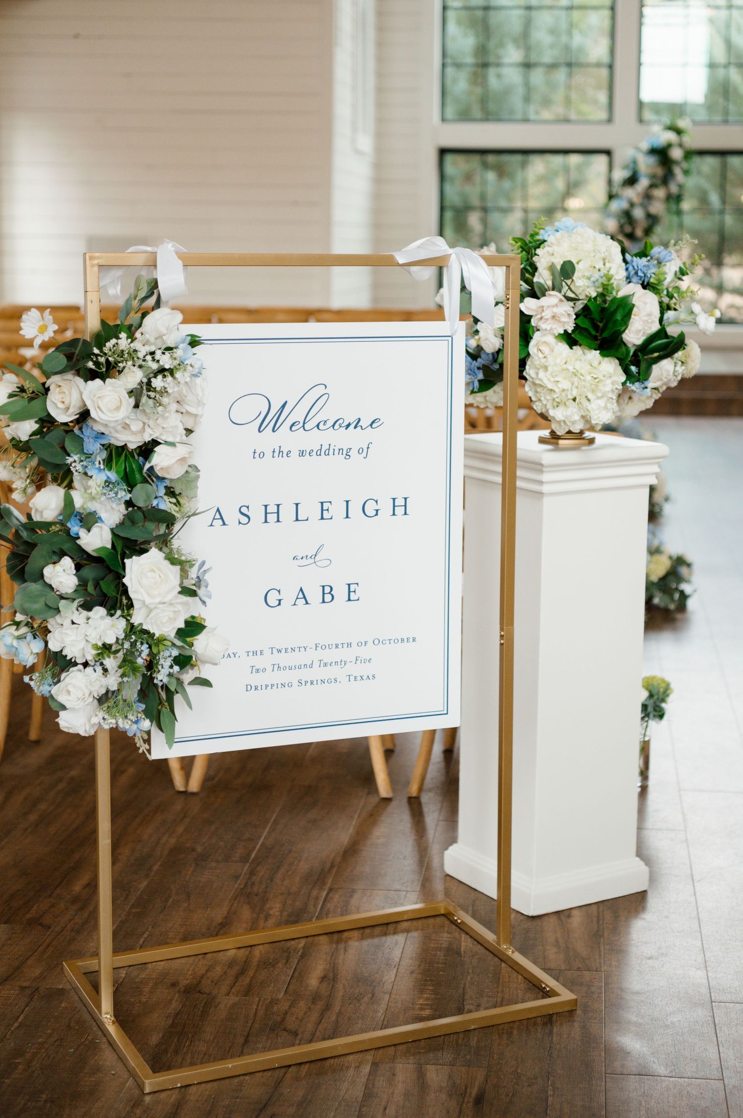 A wedding welcome sign with the names 'Ashleigh' and 'Gabe' displayed on a wooden frame, surrounded by floral arrangements.