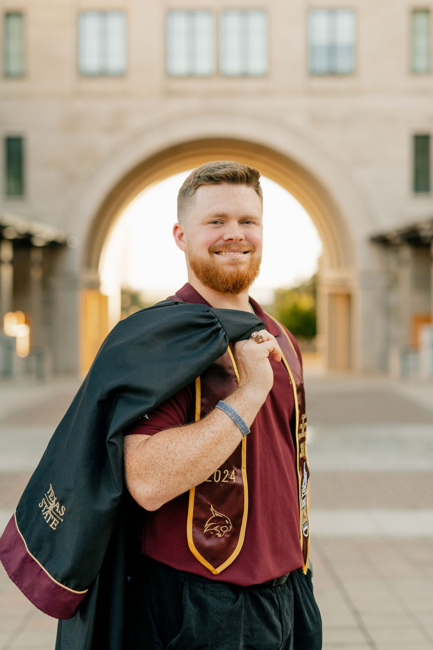 Graduate poses in front of archway wearing maroon academic regalia and black graduation stole.