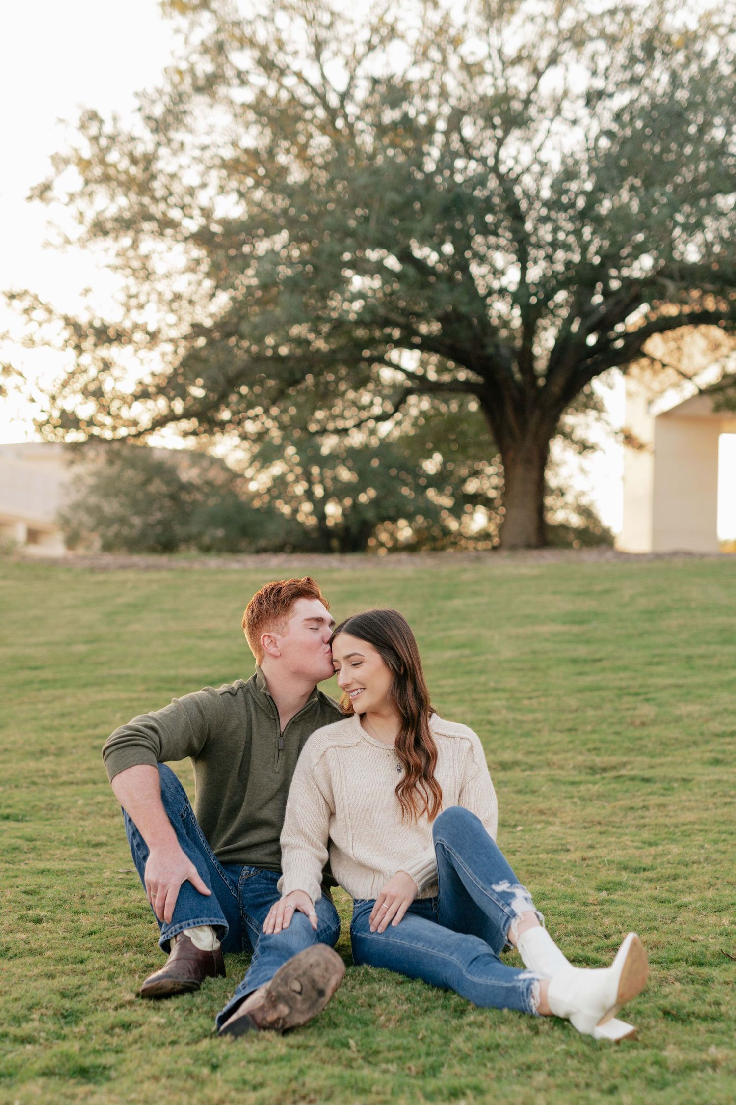 A couple sits together on a grassy hill during golden hour with a large tree silhouetted in the background.