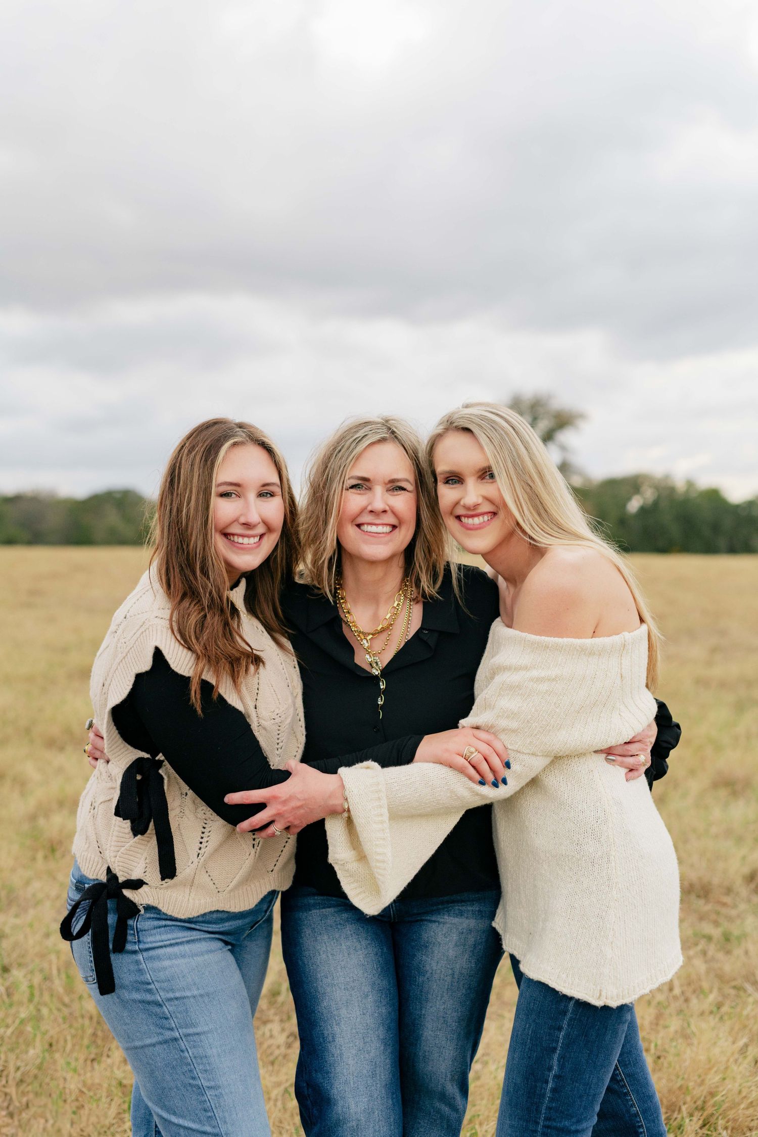 Three women in casual outfits embracing and smiling in a golden field.