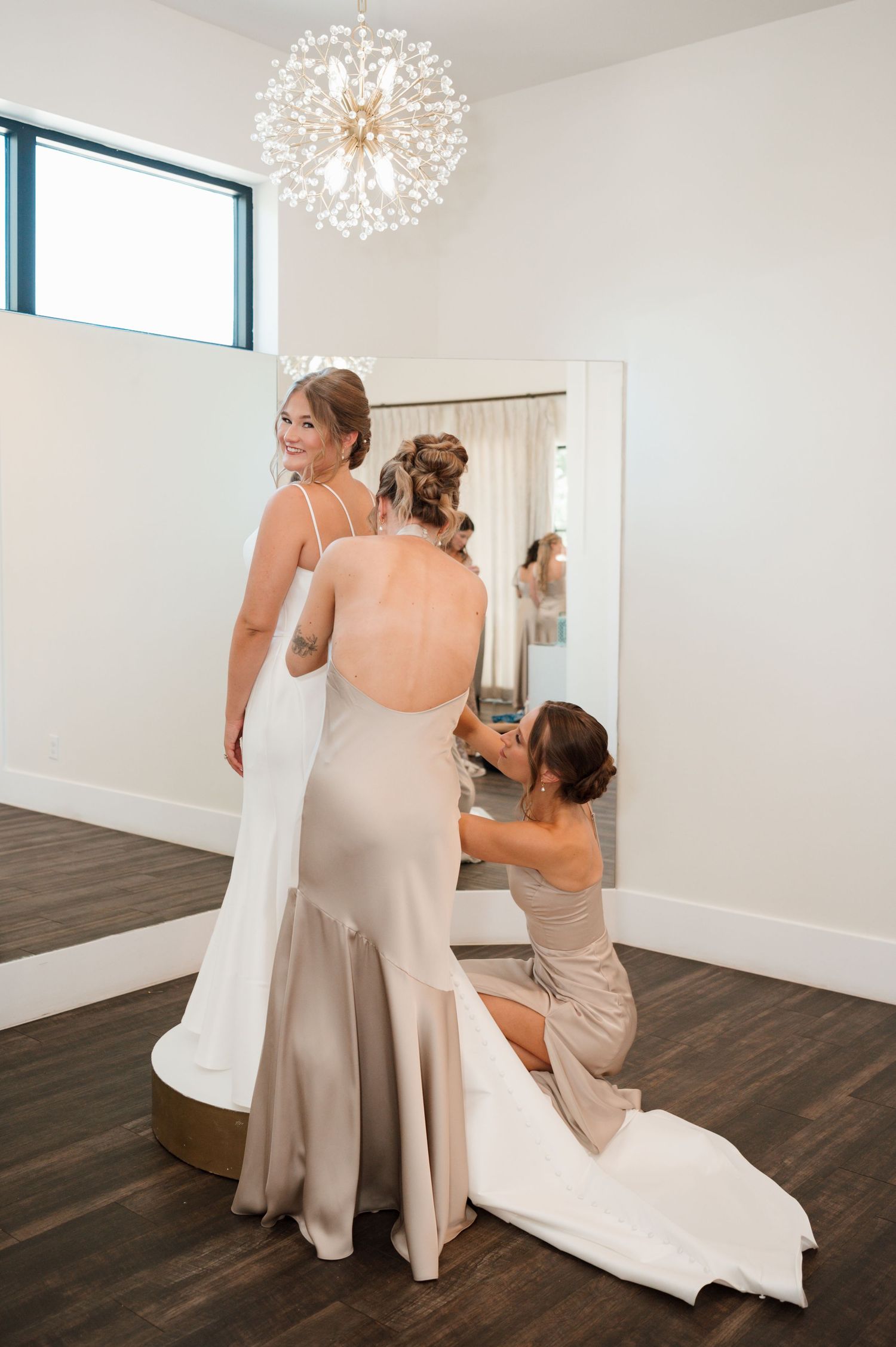 Wedding preparation scene in bright room with chandelier showing dress fitting and adjustments.