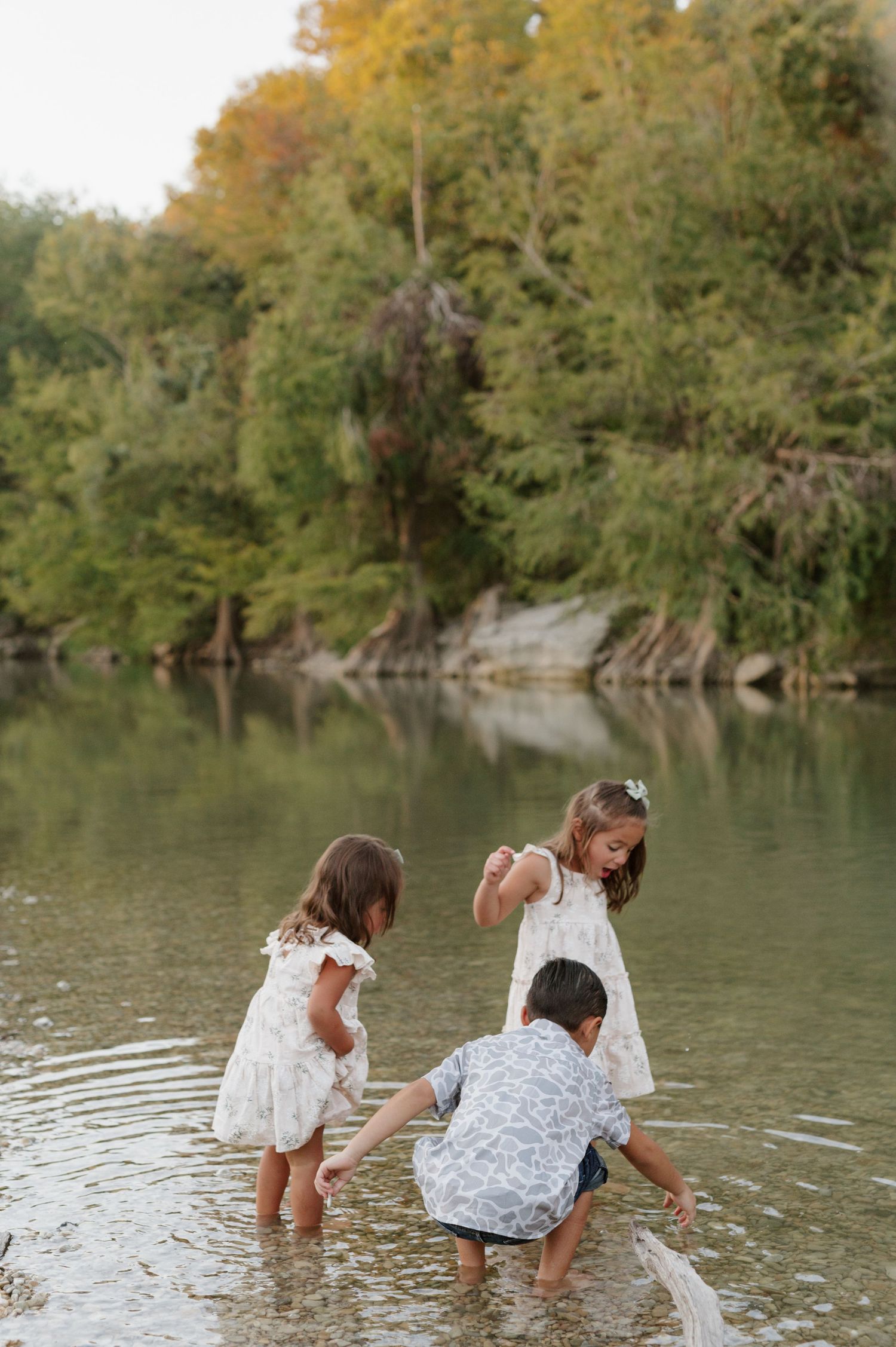 Children in white dresses splash and play together in a peaceful river surrounded by green trees on a summer day.