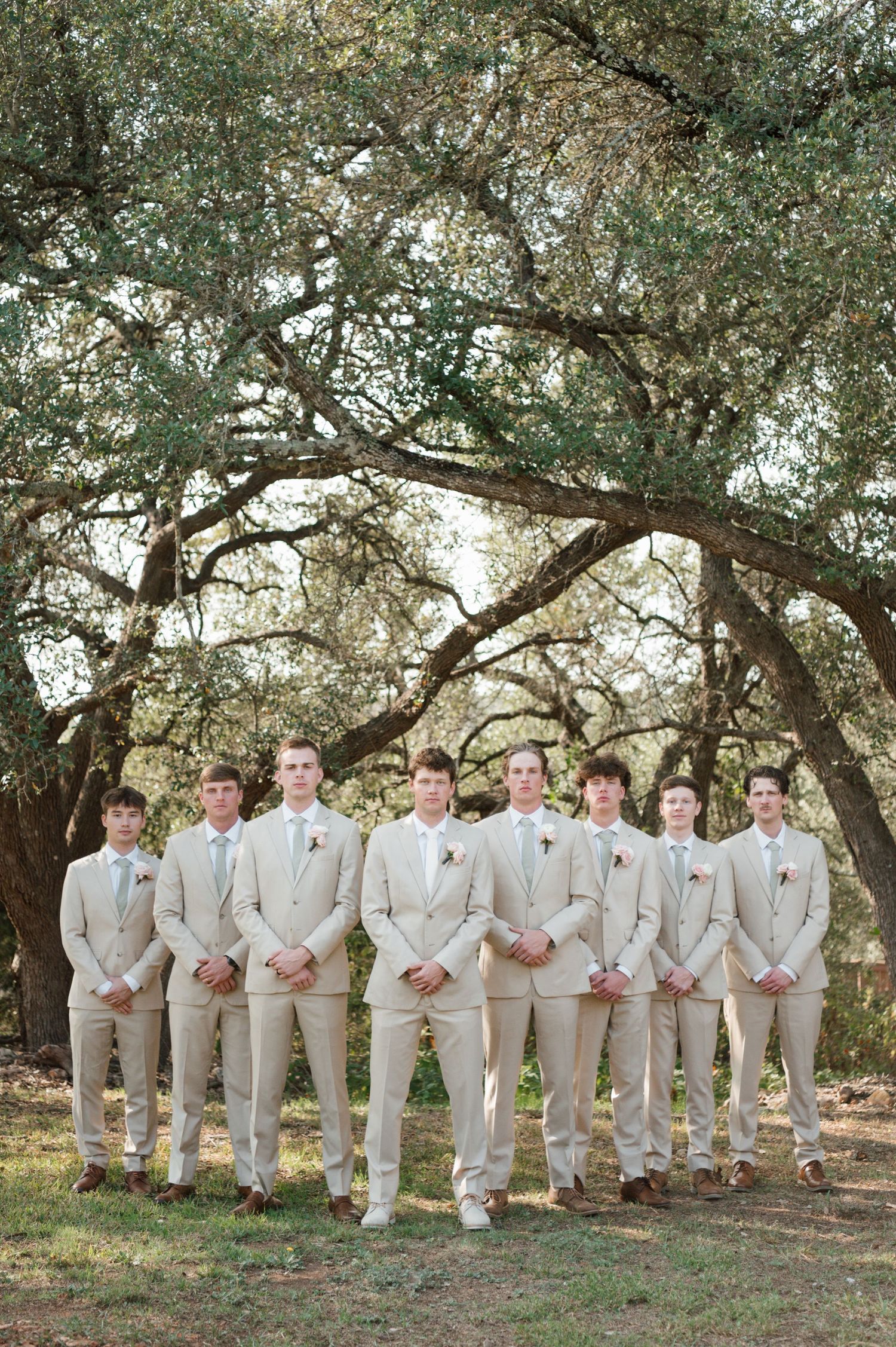Groomsmen in light beige suits pose together under oak trees at an outdoor wedding venue.