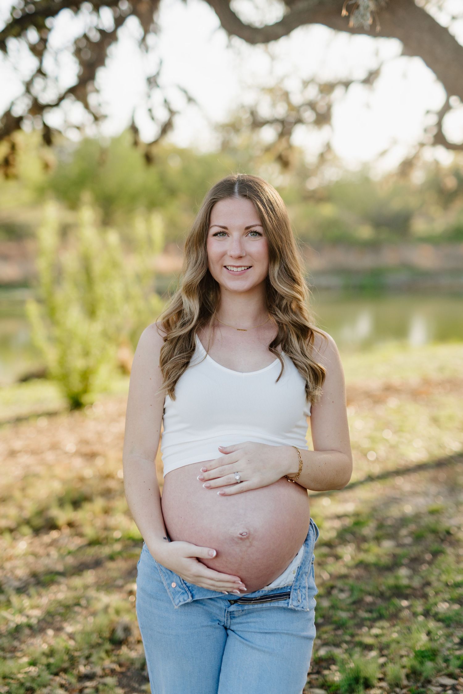 Expectant mother in white top and jeans poses outdoors near a lake during sunset maternity photography session.