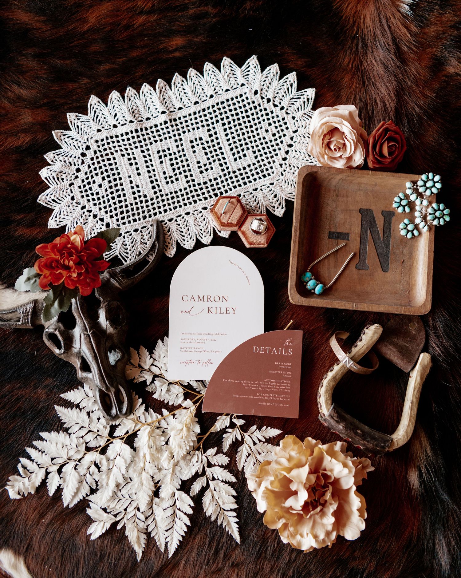 Rustic wedding decor flatlay featuring lace doily, roses, invitations, and floral elements on dark fur backdrop.