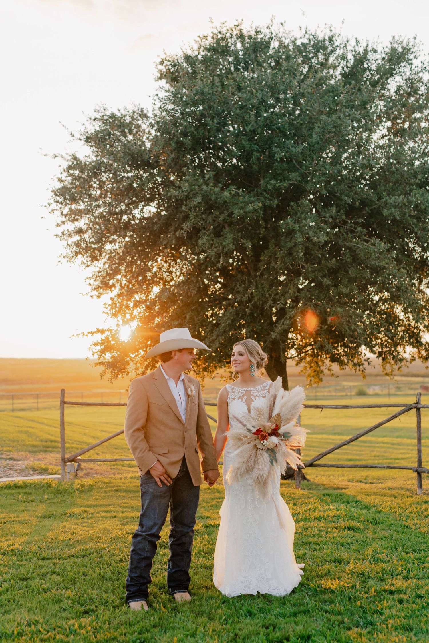 Couple in western wedding attire stands together at sunset with a majestic tree and fence silhouetted behind them.