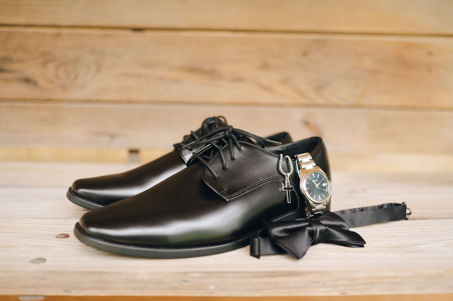 Black dress shoes with a bow tie, watch, and cufflinks on a wooden surface for a formal occasion.