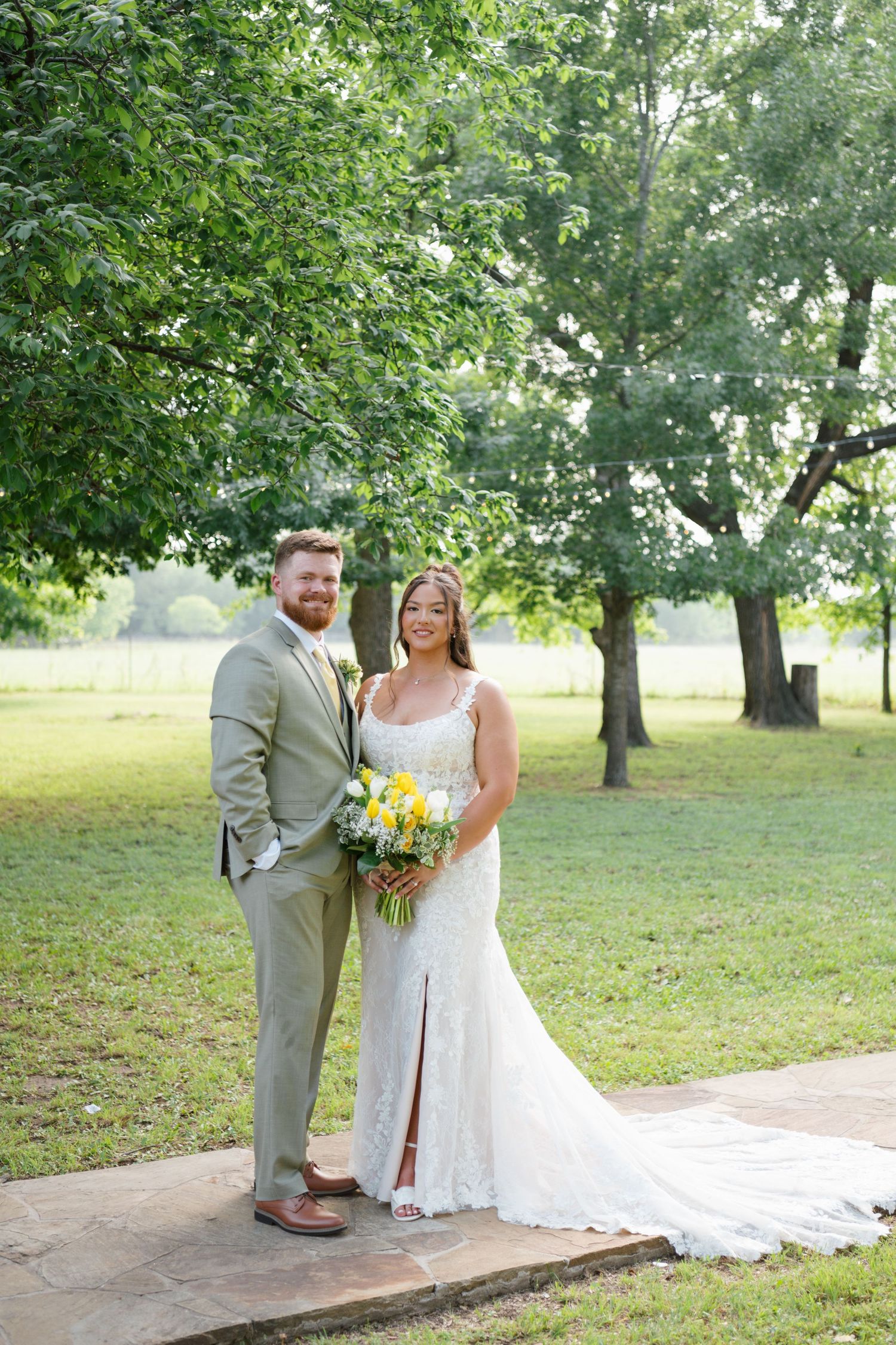 Bride and groom pose outdoors in lush green park, she holds yellow bouquet in lace gown, he wears sage suit.