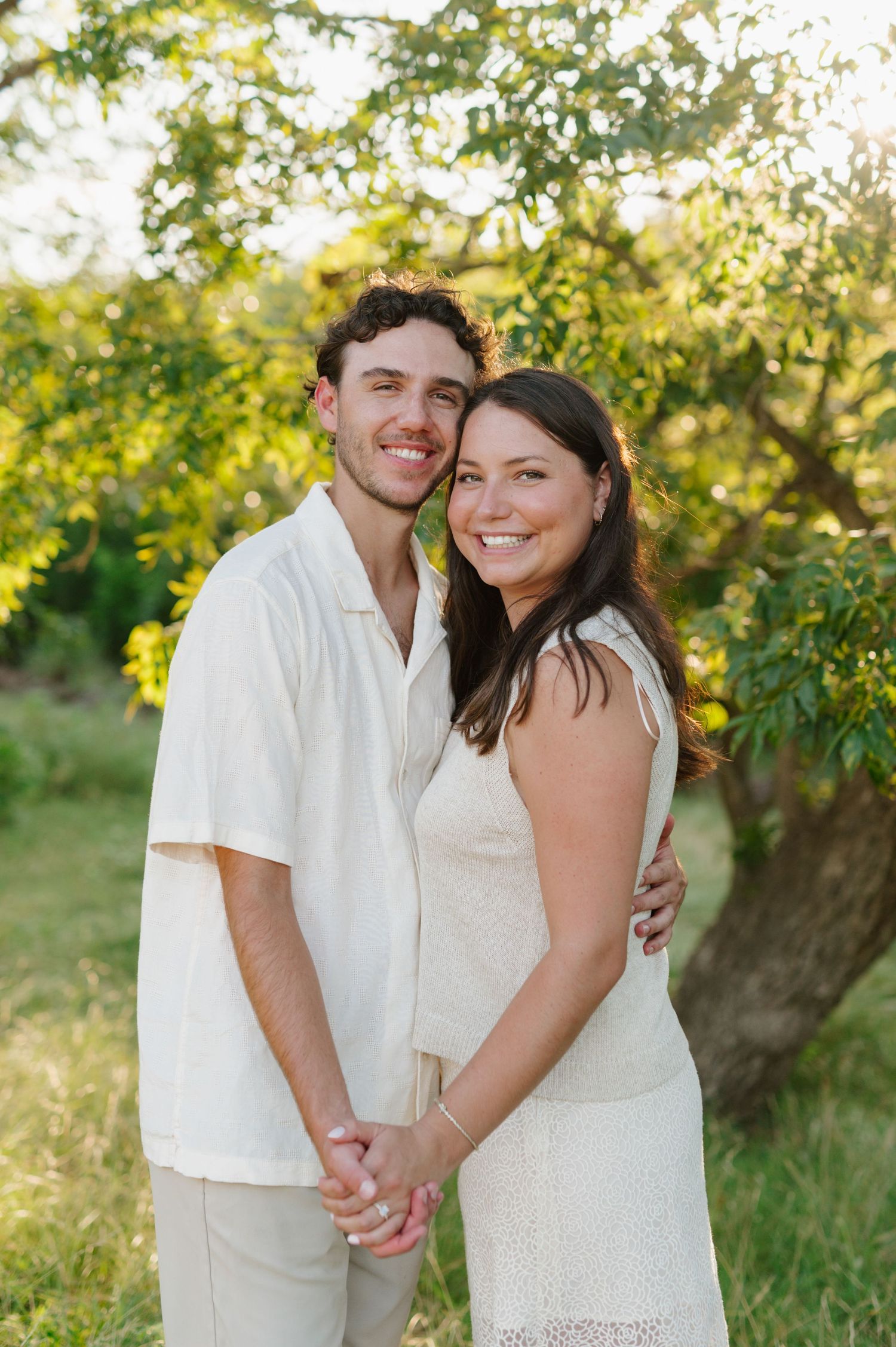 A couple in white outfits sharing loving embraces against a sunlit natural backdrop.