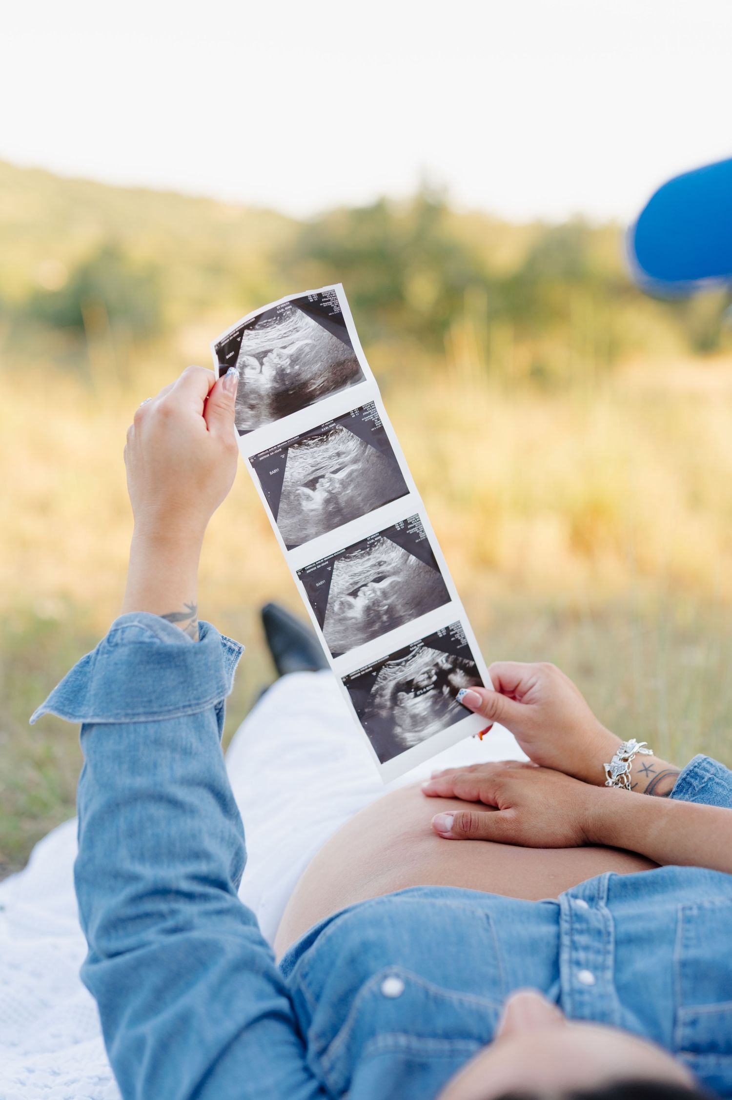 A person lying down in denim jeans while holding ultrasound photos in a grassy outdoor setting.