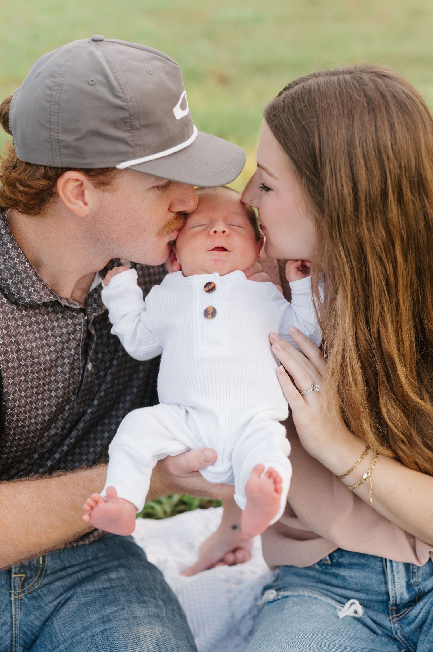 Parents kiss their newborn baby dressed in white while sitting together outdoors.