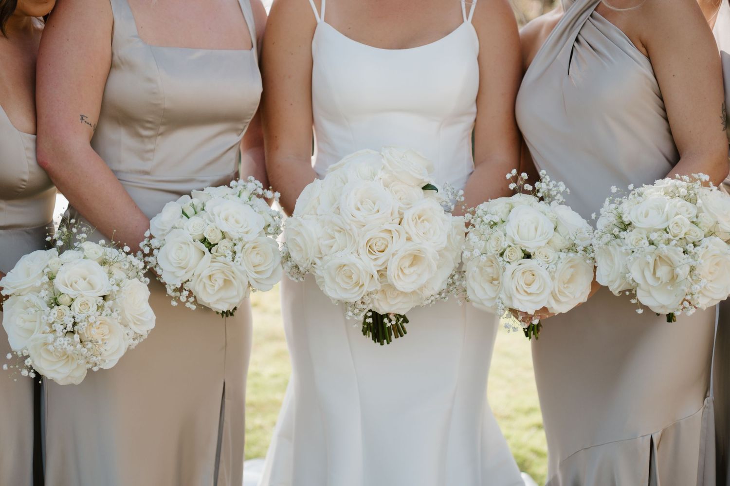 Bridesmaids in beige dresses stand together holding elegant white peony and rose bouquets at an outdoor wedding.