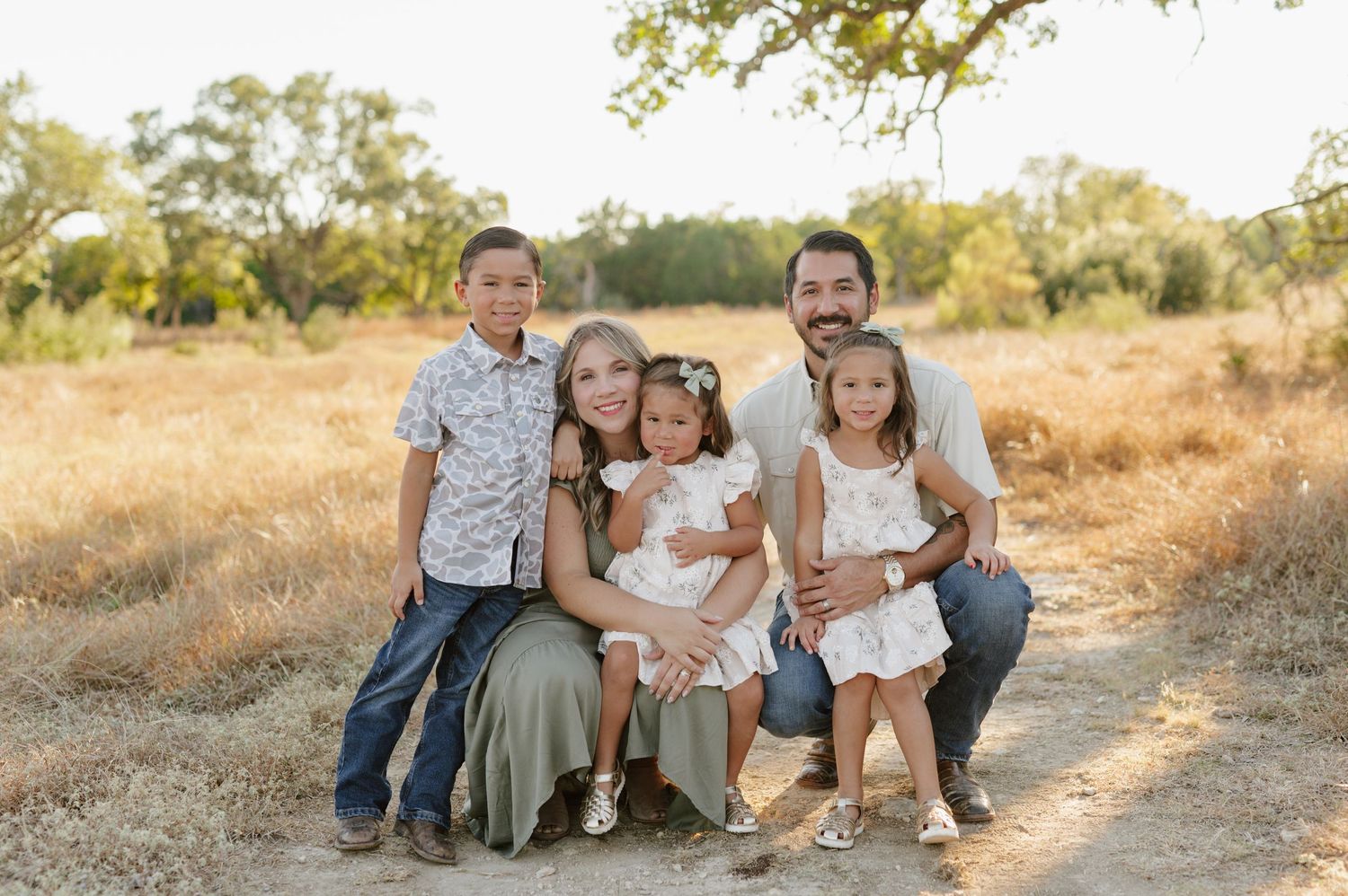 A family group poses together outdoors on a dirt path with golden grass and trees in the background.