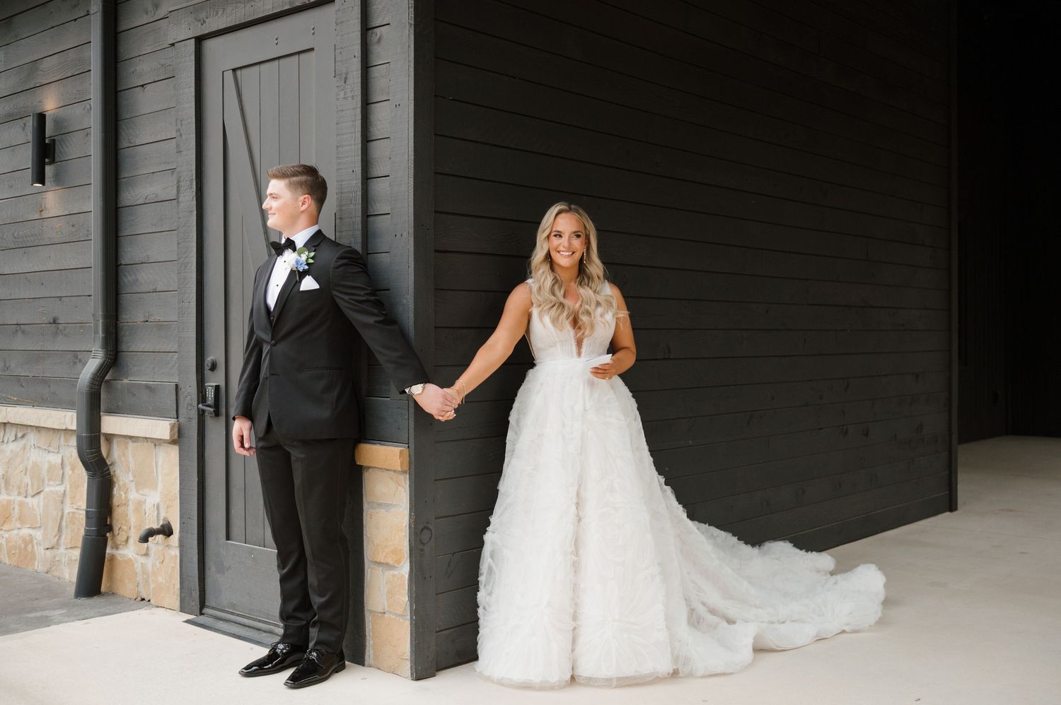 A bride in a white lace dress and a groom in a black tuxedo standing together outside a wooden building.