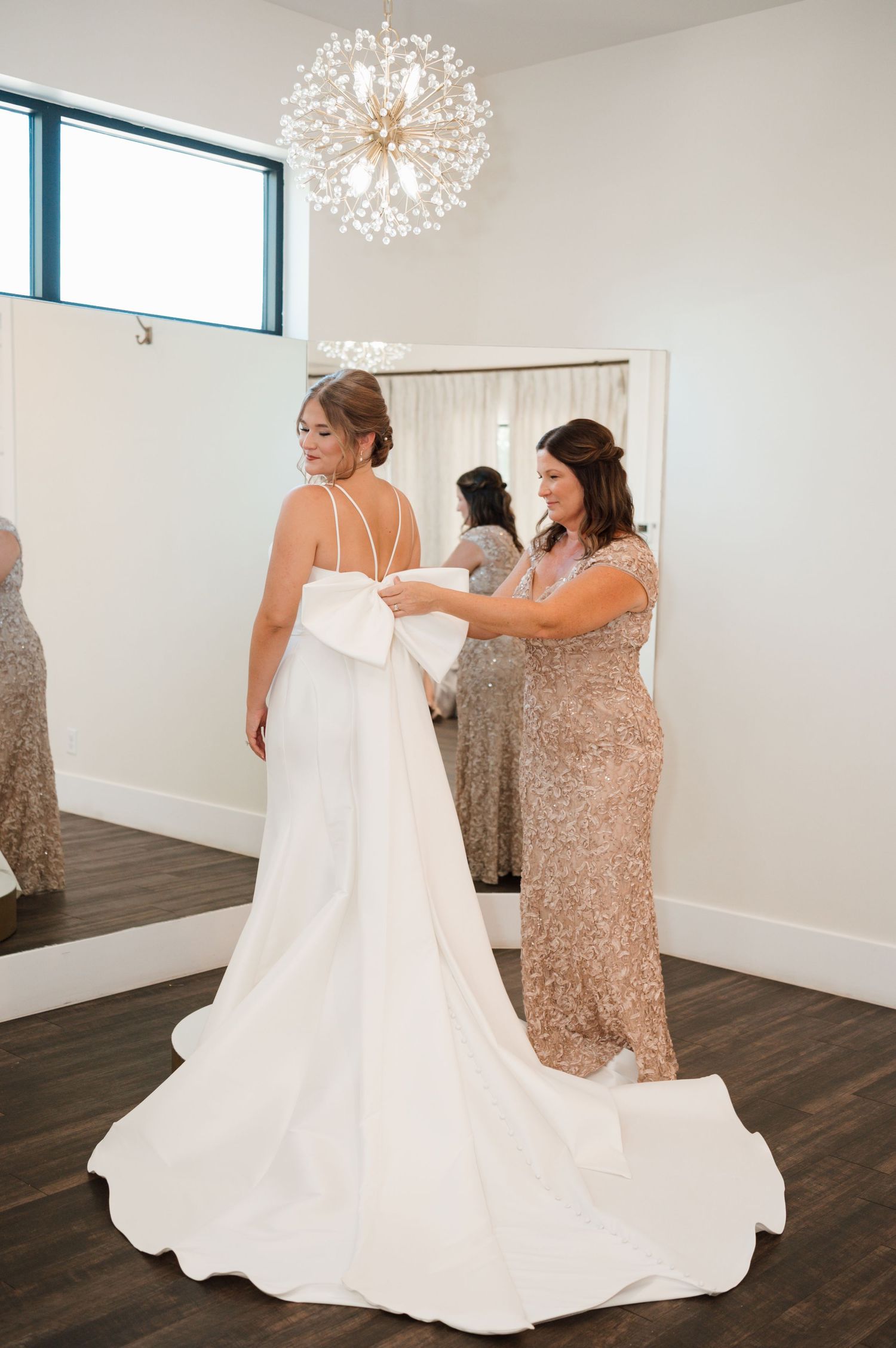 Wedding preparations showing the flowing white dress being adjusted in a bright room with chandeliers.