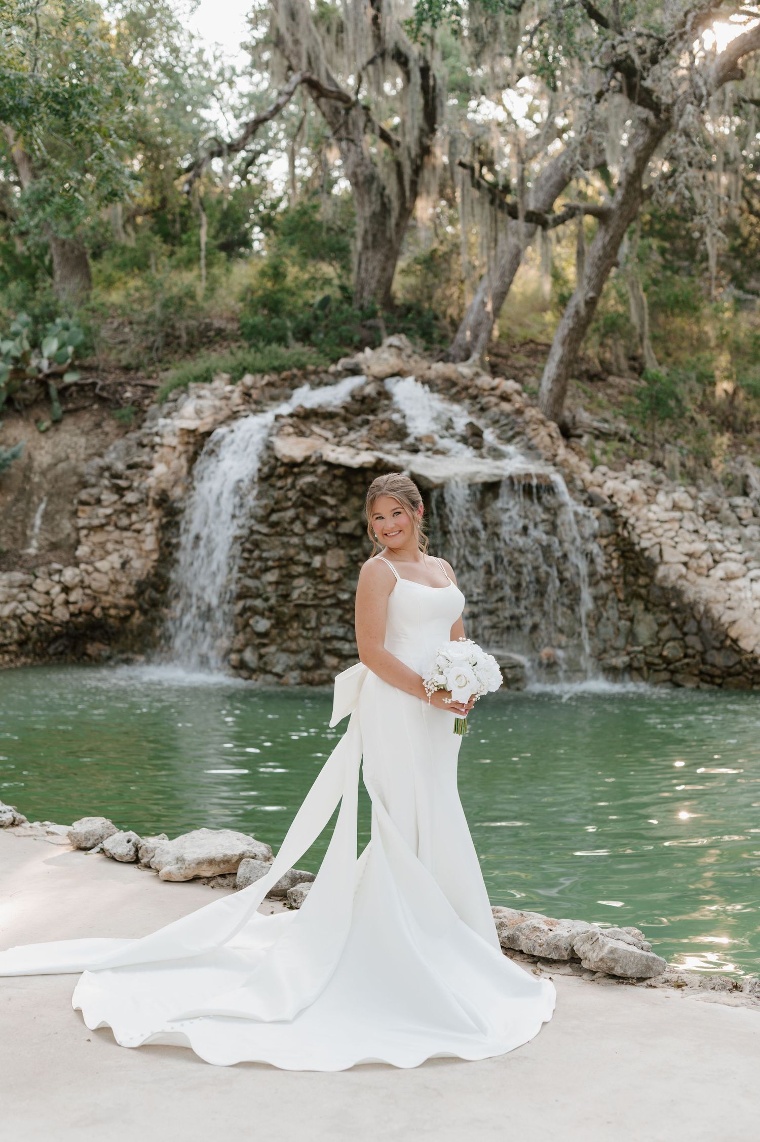 An elegant wedding dress displayed against a scenic waterfall and turquoise pool backdrop in a natural setting.