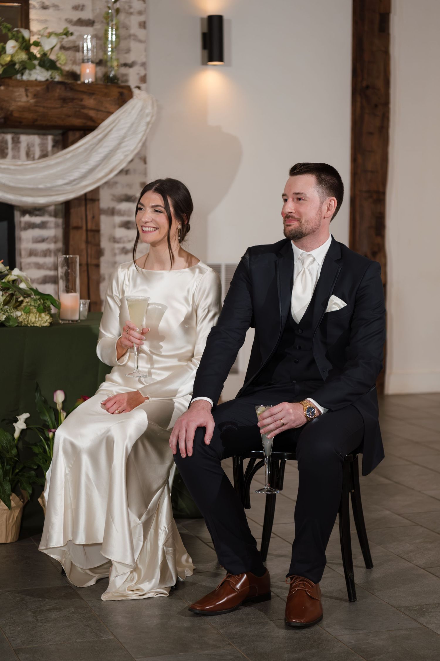 Elegant wedding couple seated in rustic venue with exposed brick walls and white drapery.