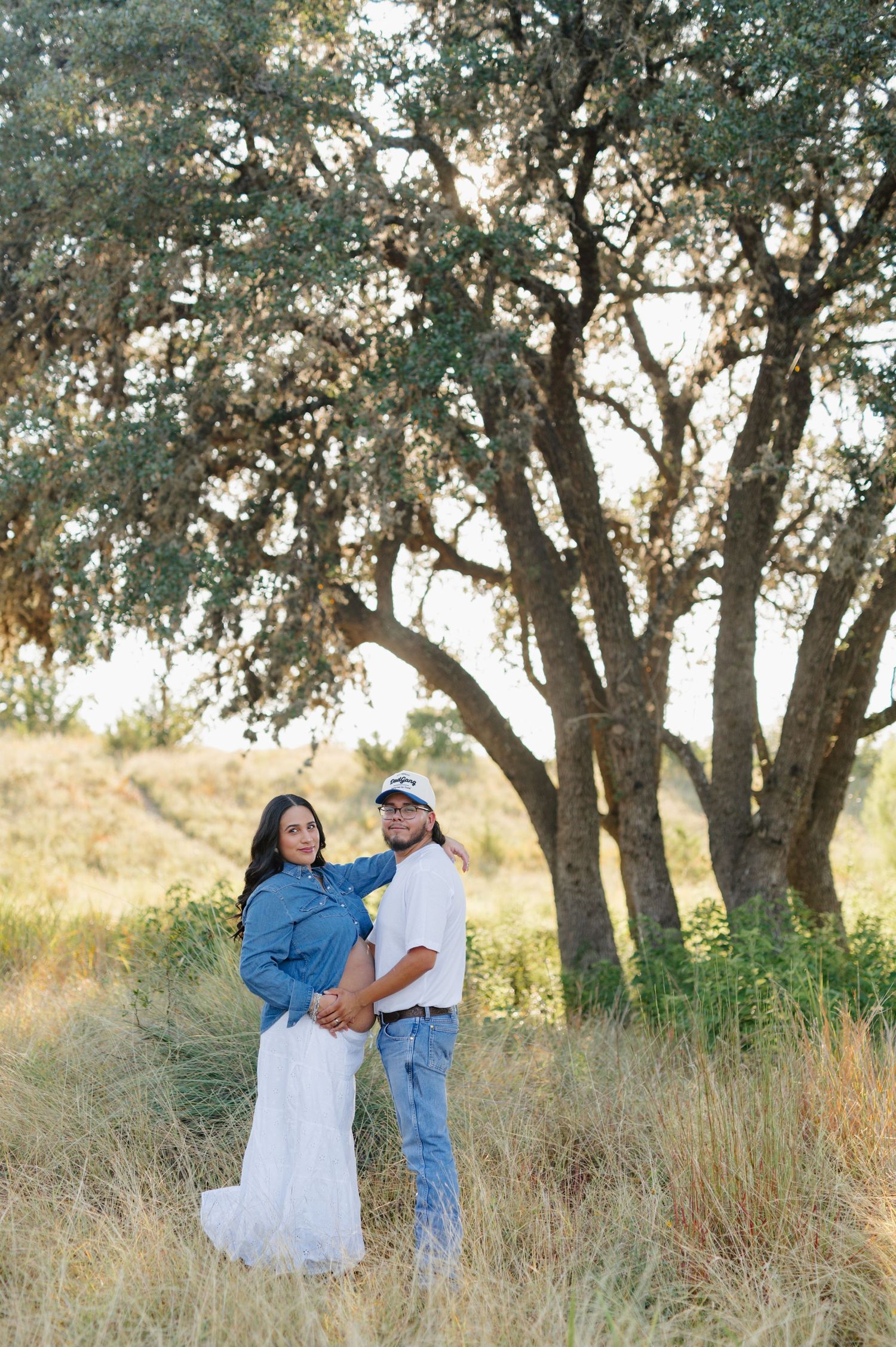 Couple shares intimate moment among oak trees in a serene outdoor field setting.