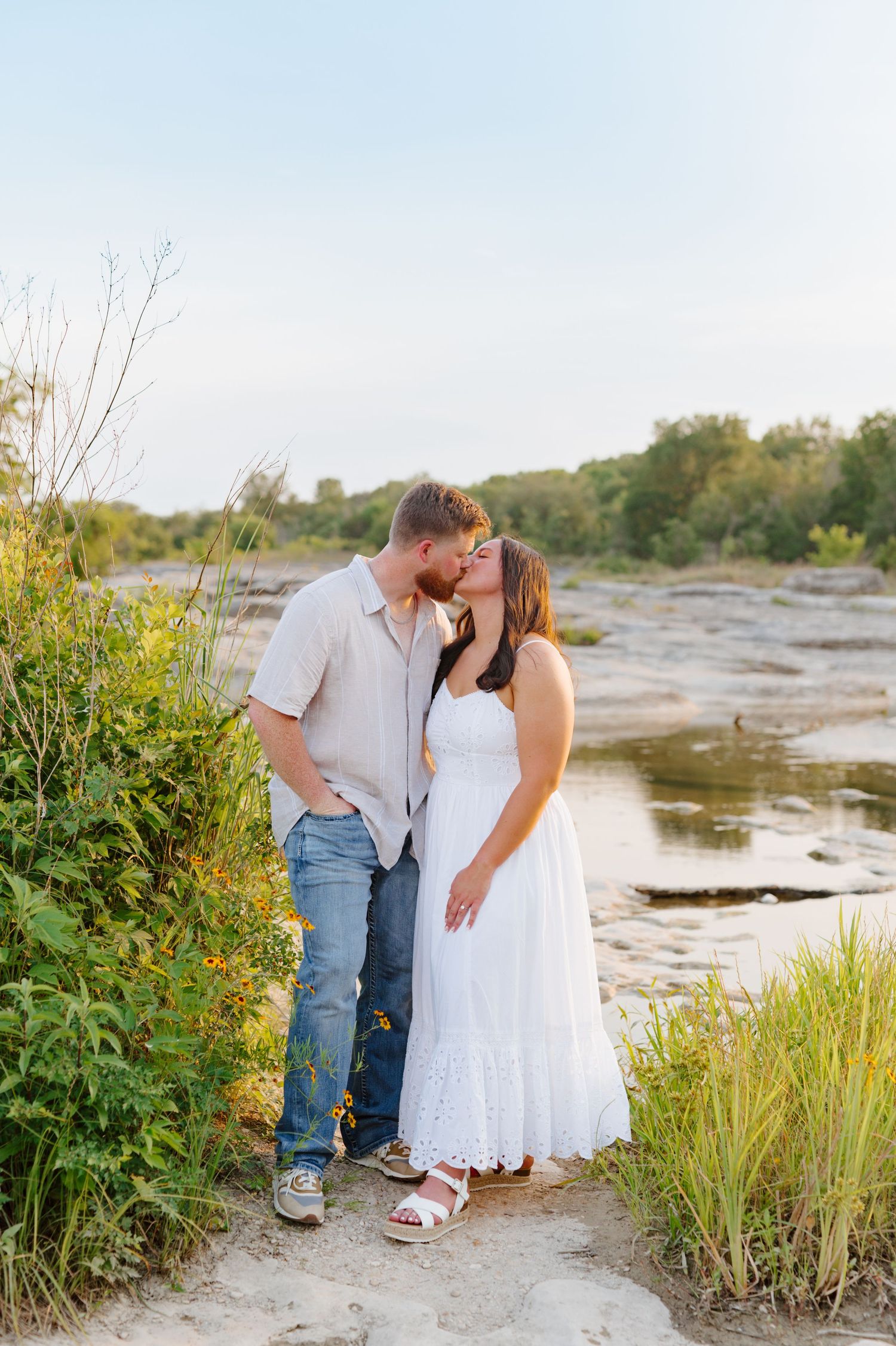 A couple shares a romantic moment on a riverbank surrounded by wild grasses in golden evening sunlight.