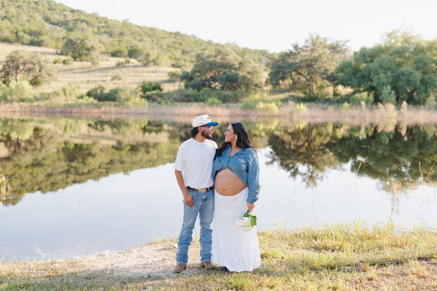 Couple's maternity photos taken by a serene lake during golden hour with hills in the backdrop.