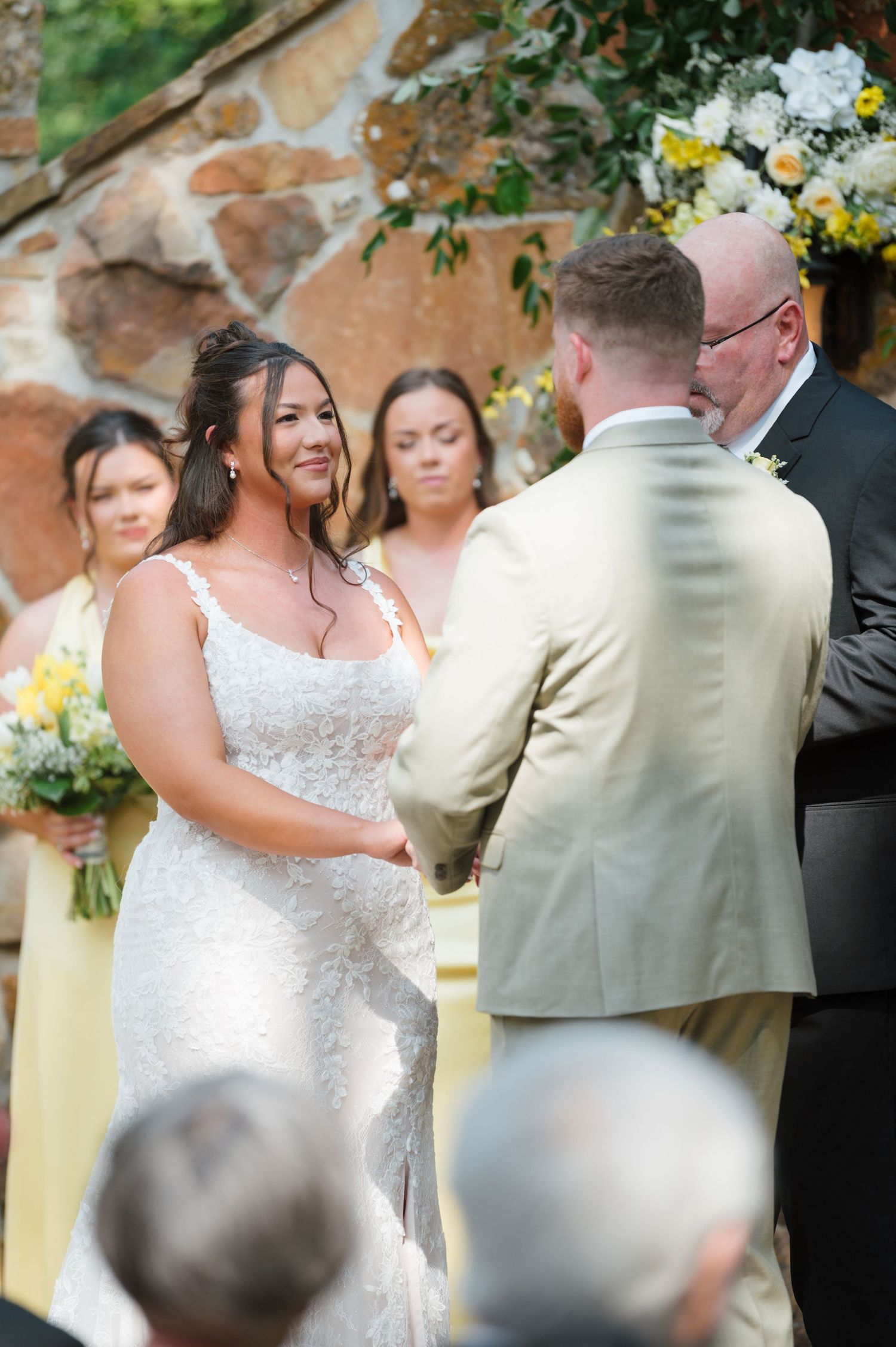 Smiling bride in lace gown exchanging vows with groom in tan suit at outdoor stone ceremony.