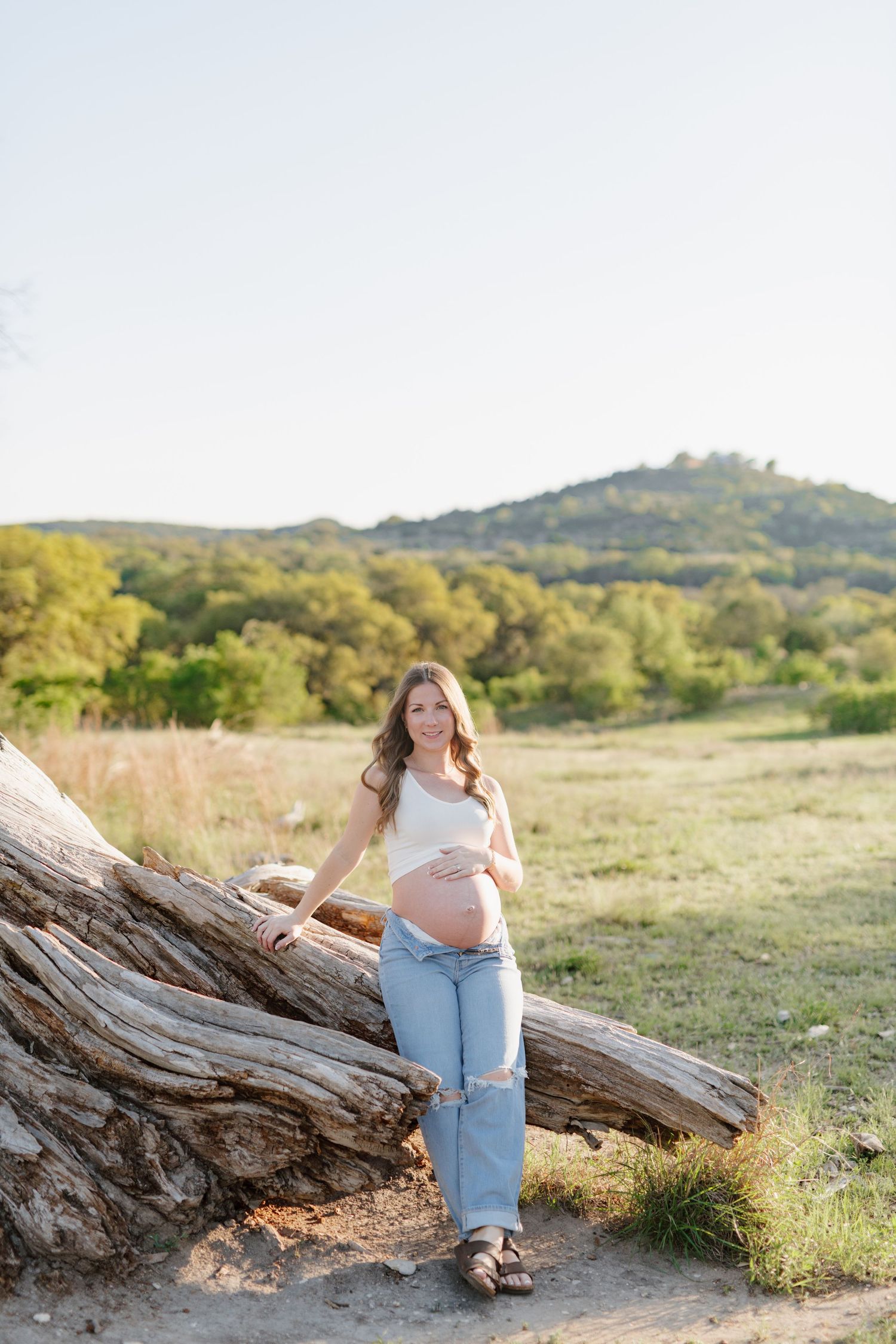 Outdoor maternity photo in golden sunlight with casual jeans and white top against rustic fallen tree trunk.