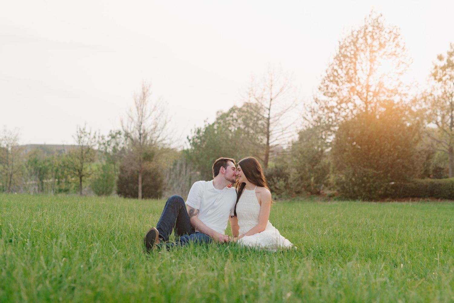A romantic couple sits together in a sunlit meadow surrounded by green grass and trees.