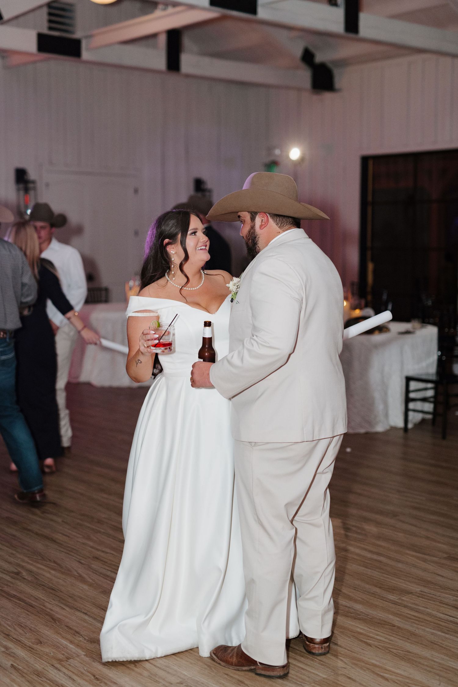 A couple shares a tender moment on the dance floor at a rustic wedding reception, both holding drinks.