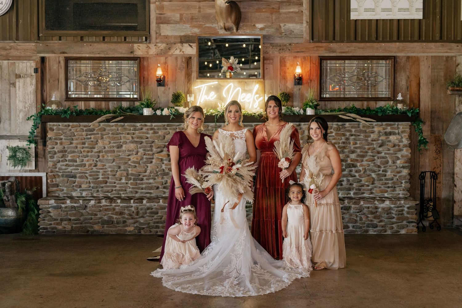 Wedding party poses in front of stone bar wall with bridesmaids in burgundy and neutral dresses.