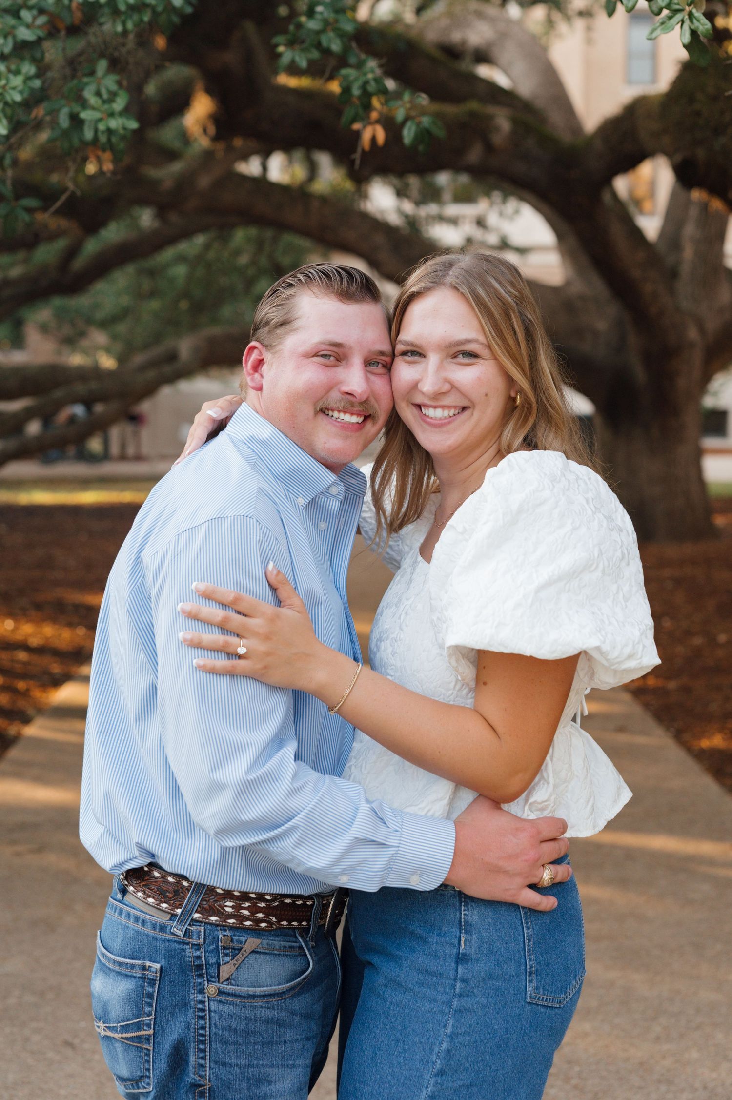 A couple in jeans and light blue and white tops embrace and laugh together under large oak trees.