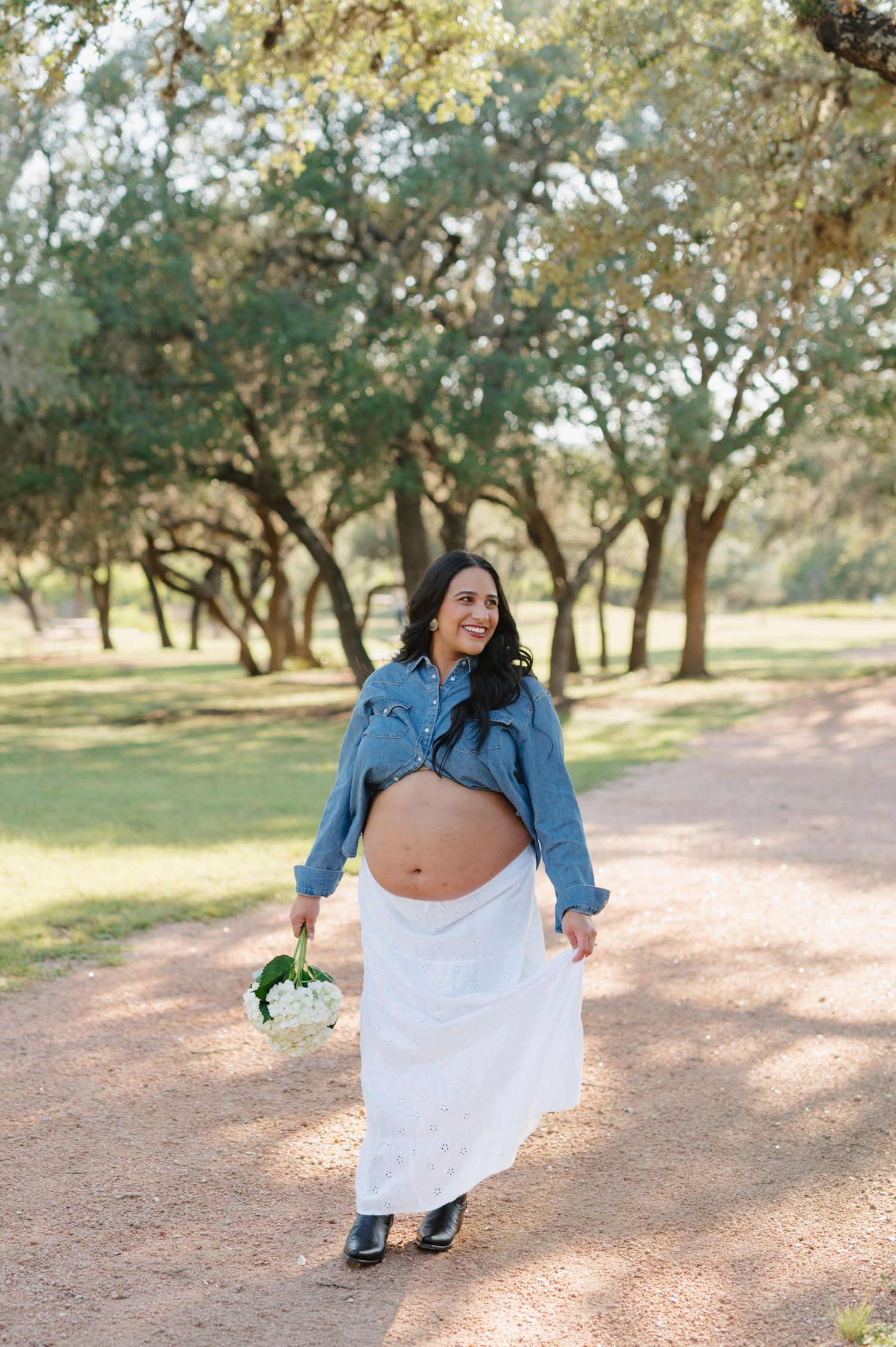 An expectant mother in a denim jacket and white skirt poses on a sunlit path surrounded by trees.
