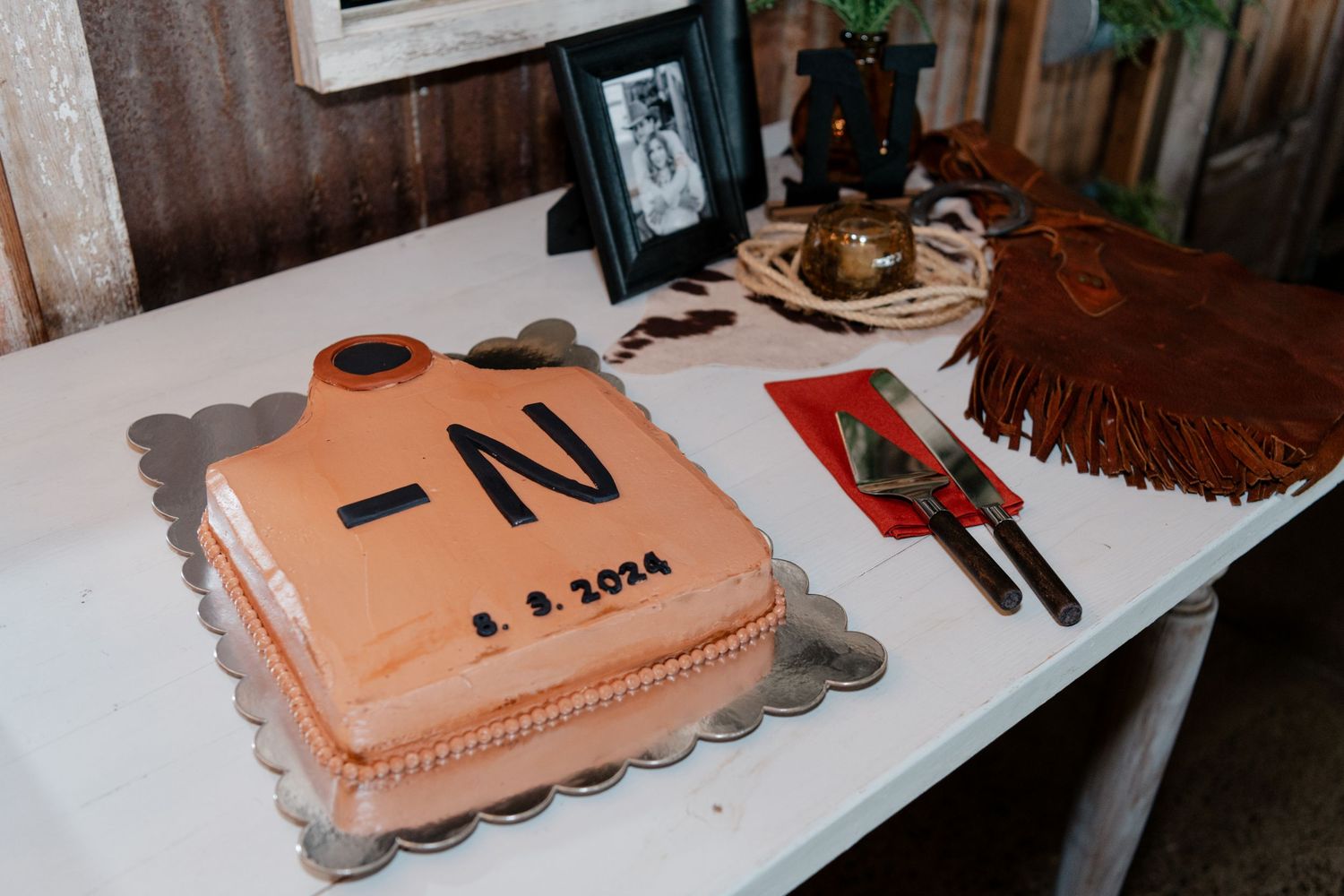 Pink square wedding cake with letter N decoration displayed on white table with serving utensils.