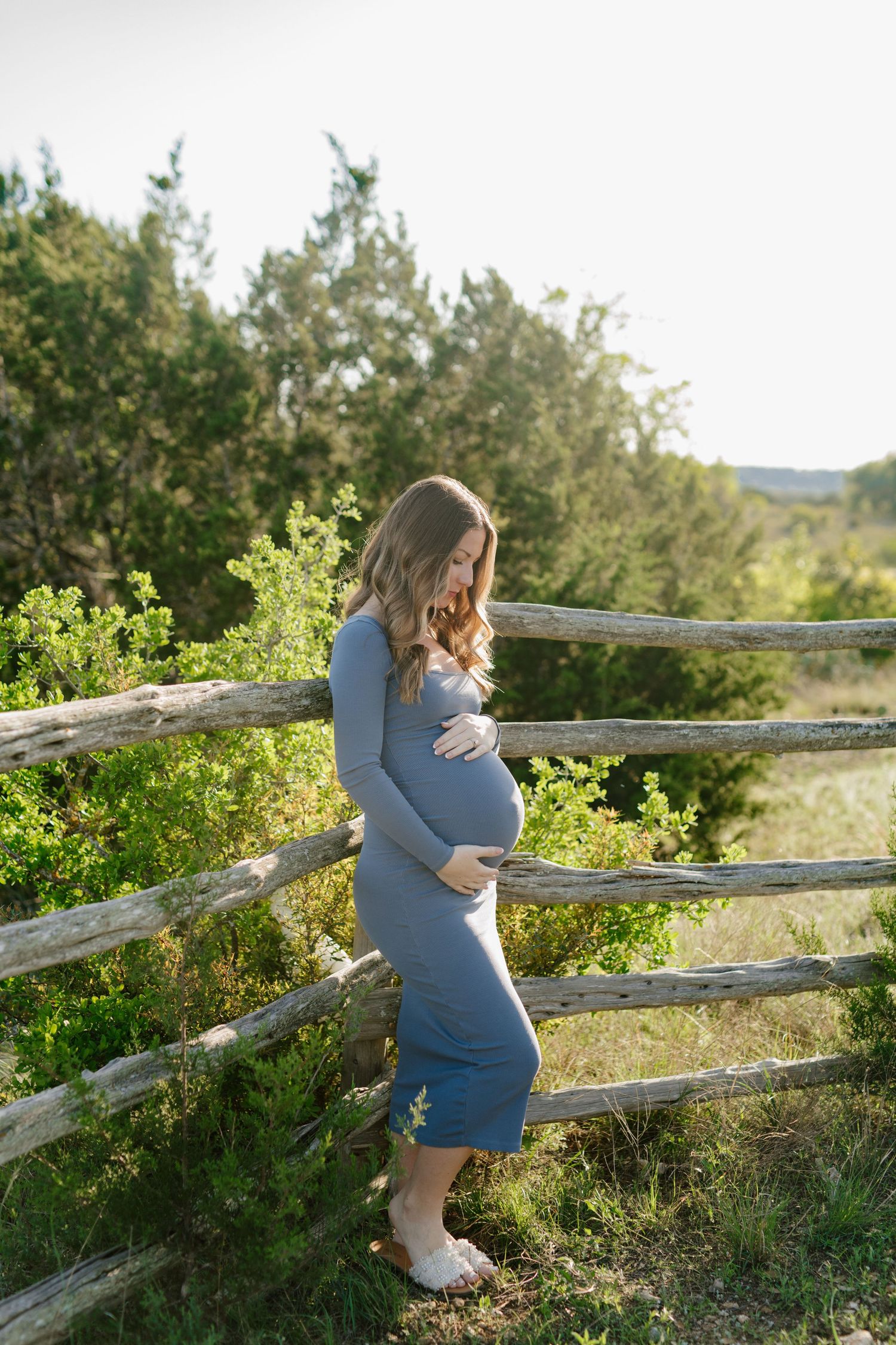 Maternity photo of an expecting mother in a blue dress standing by a rustic wooden fence surrounded by greenery at sunset.