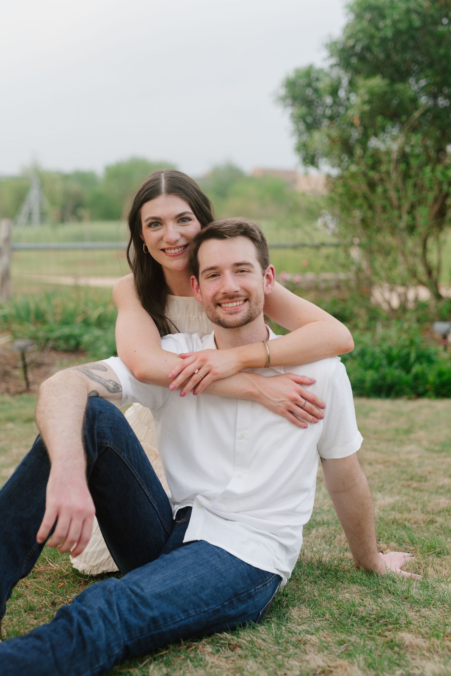 A couple sits on grass outdoors sharing an intimate embrace in a natural setting with trees in the background.