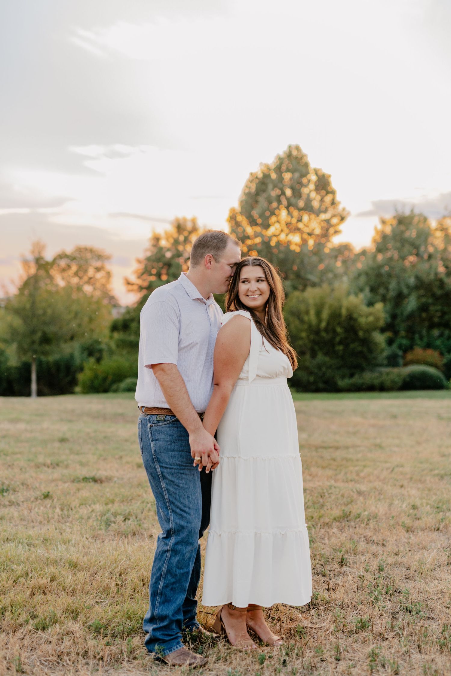 Couple holds hands while posing in a field during sunset with glowing trees in the background.