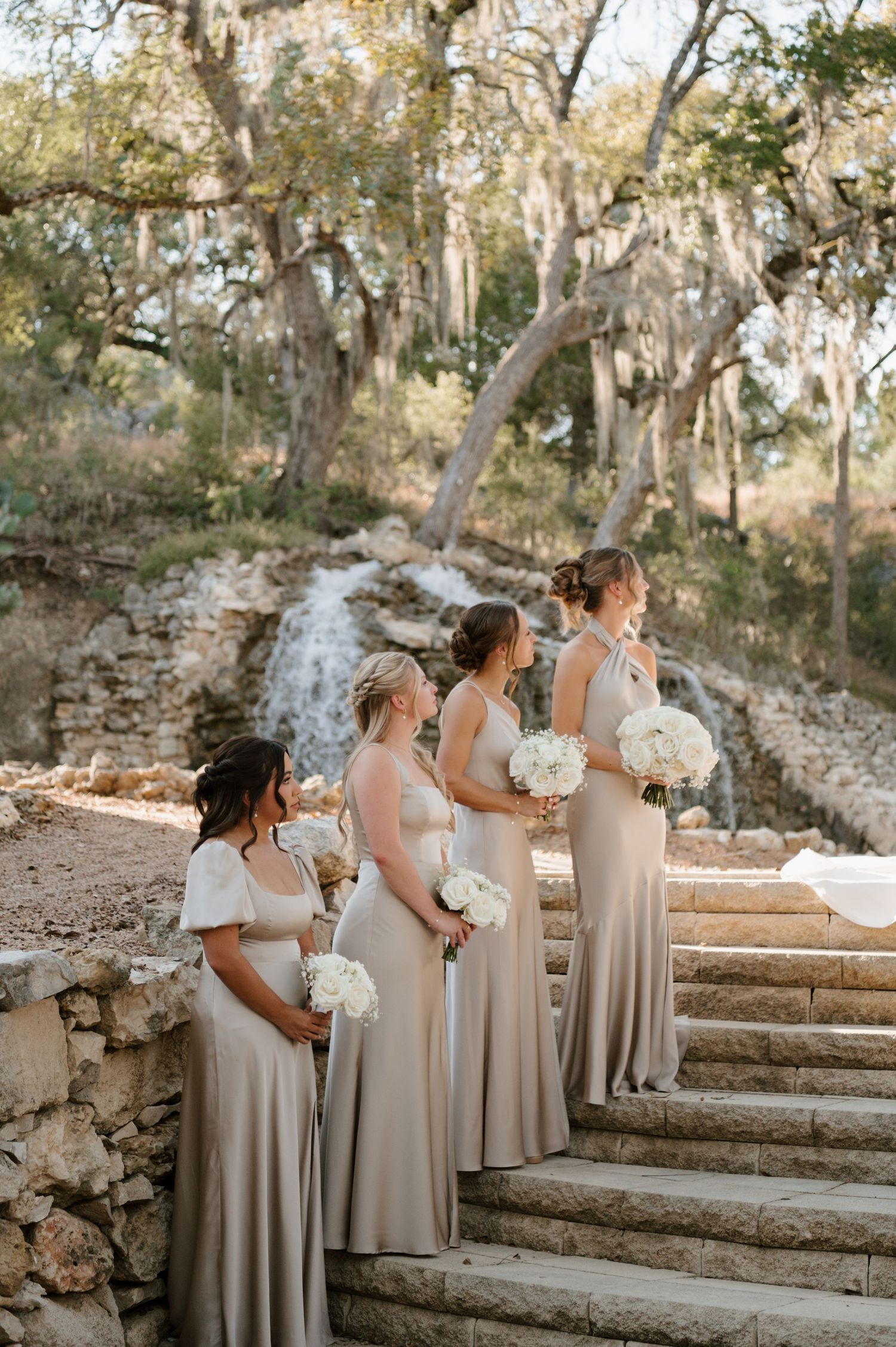 Bridesmaids in neutral dresses standing on rustic stone steps during outdoor ceremony.