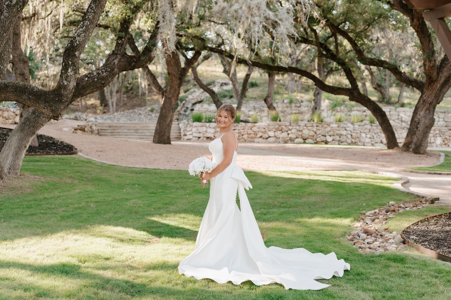 A bride in a flowing white dress twirls gracefully in a garden setting surrounded by trees and natural light.