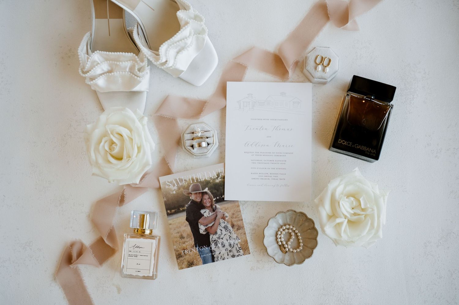 Elegant wedding details flatlay with white roses, perfume bottles, and delicate paper invitations on neutral background.