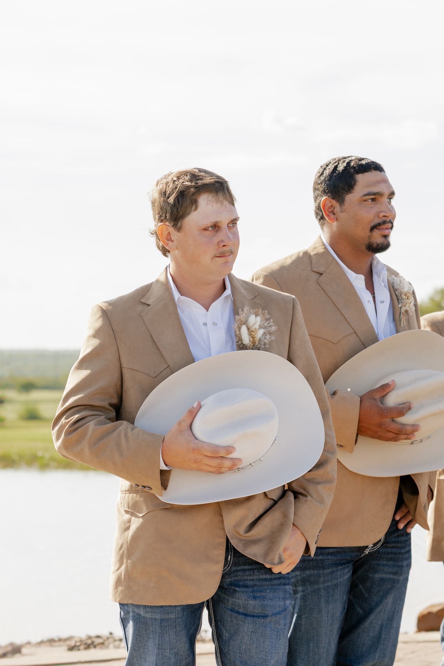 Two groomsmen in tan blazers holding cowboy hats against bright background.