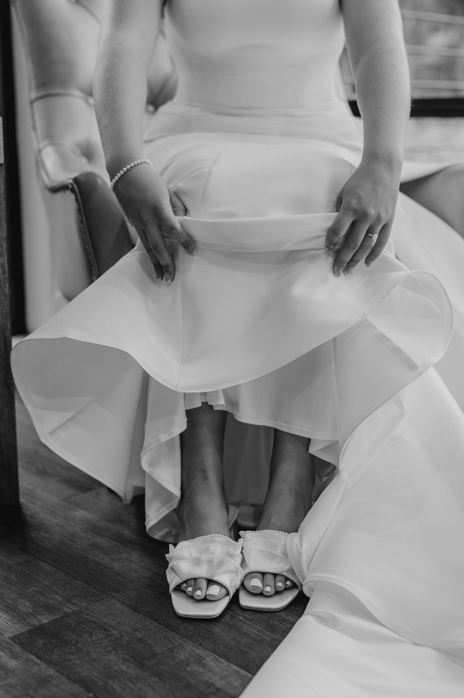 Close-up of white wedding dress hem and bridal shoes on dark hardwood floor.