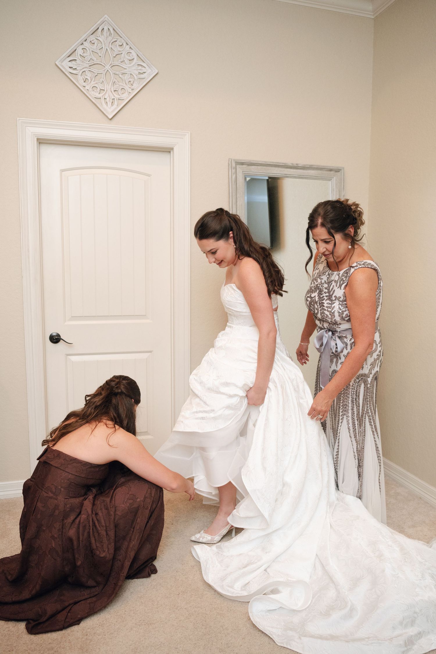 Bride in strapless white gown gets assistance from two bridesmaids adjusting her dress and shoes.