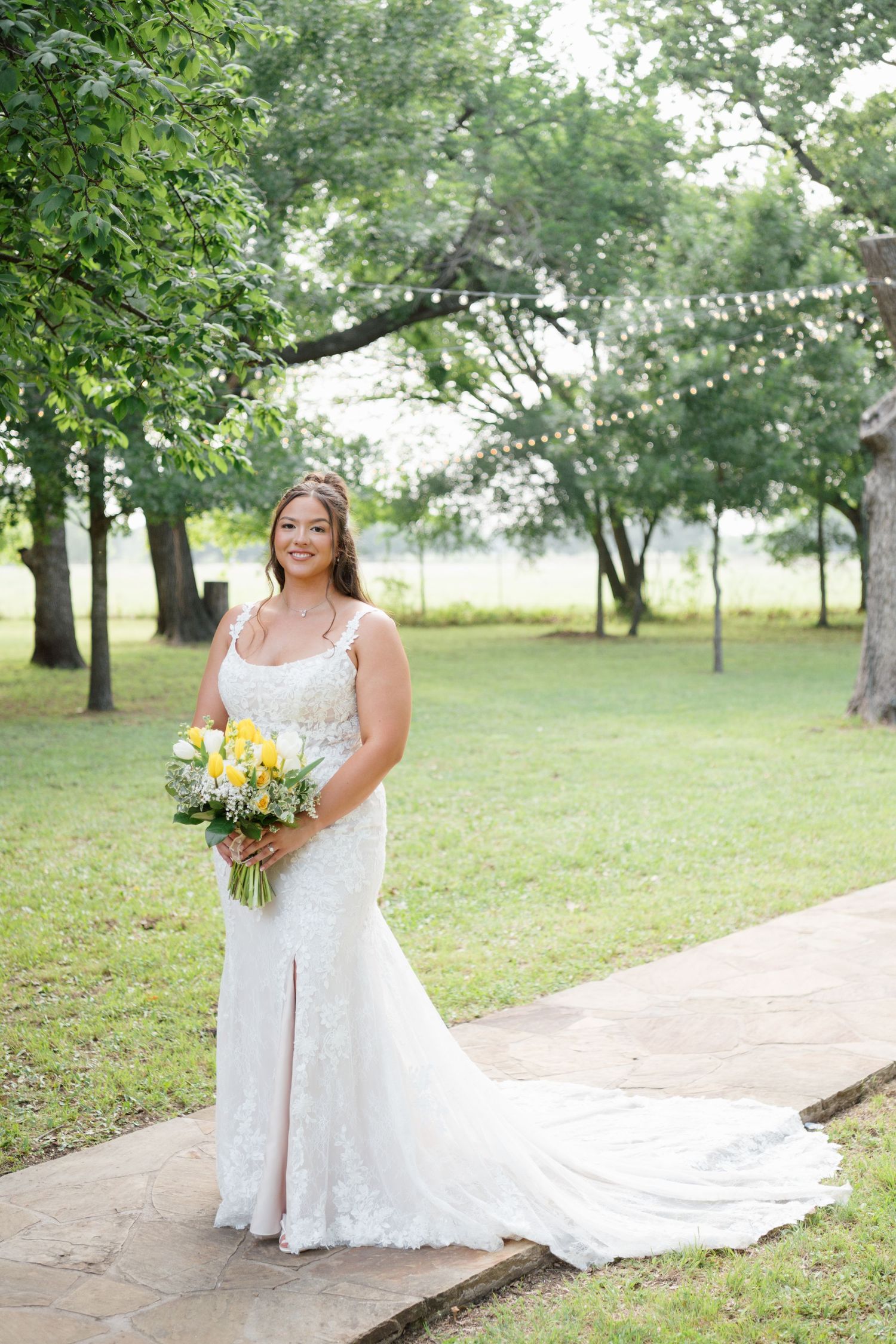 Bride in lace mermaid gown holding yellow bouquet poses on garden path surrounded by trees.