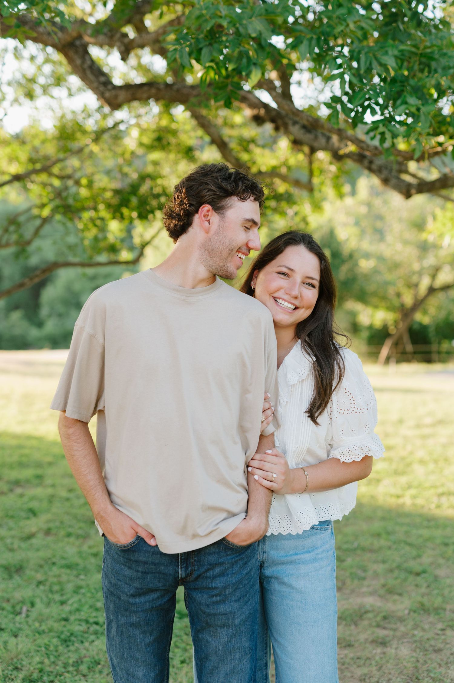 Couple in casual attire sharing intimate moments under green trees in a sunlit field.