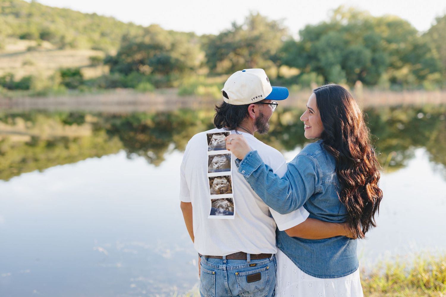 Expectant parents share tender moment outdoors while displaying sonogram photo near waterfront at golden hour.