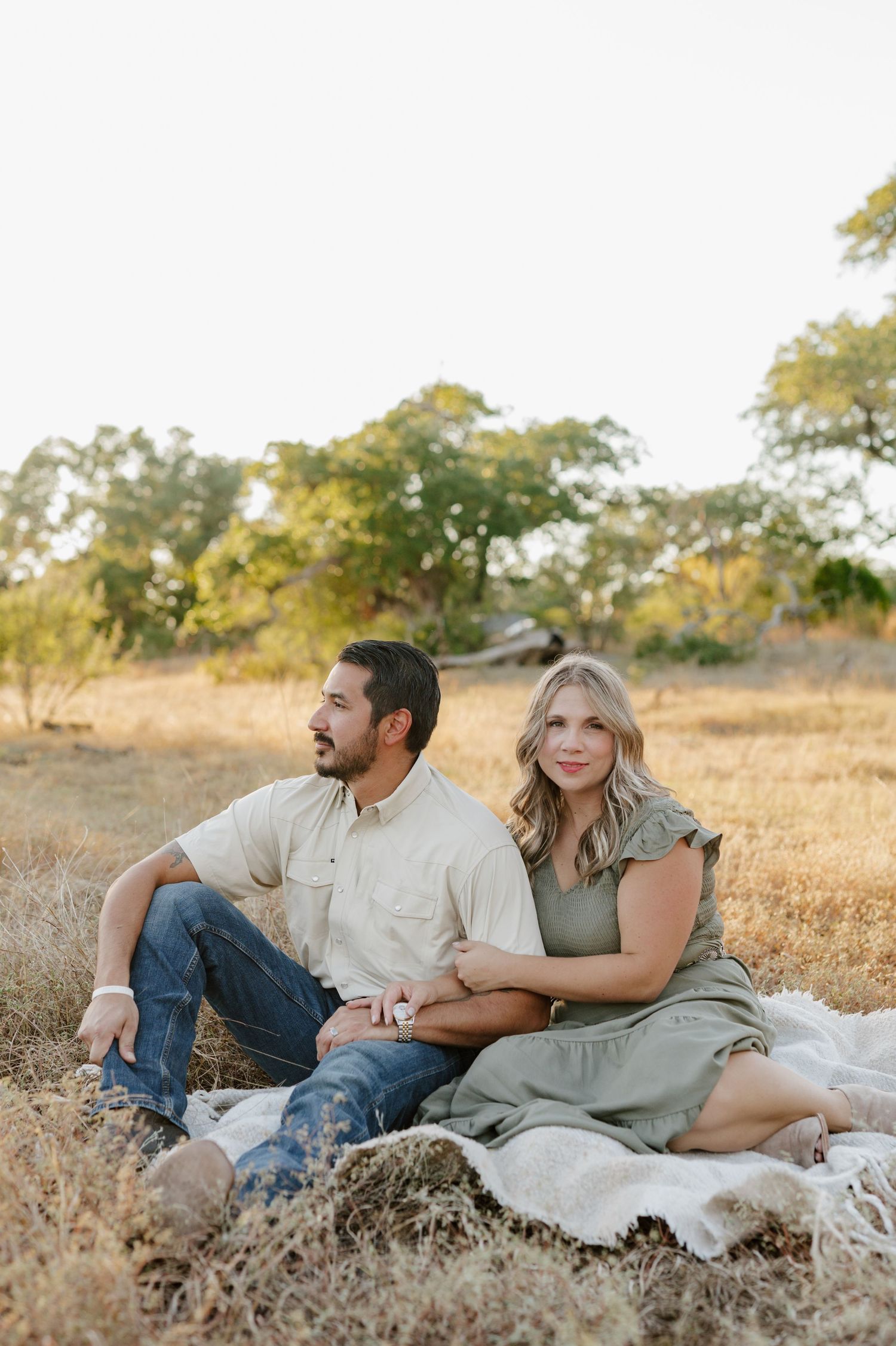 A couple relaxes together on a blanket in a golden field during sunset.