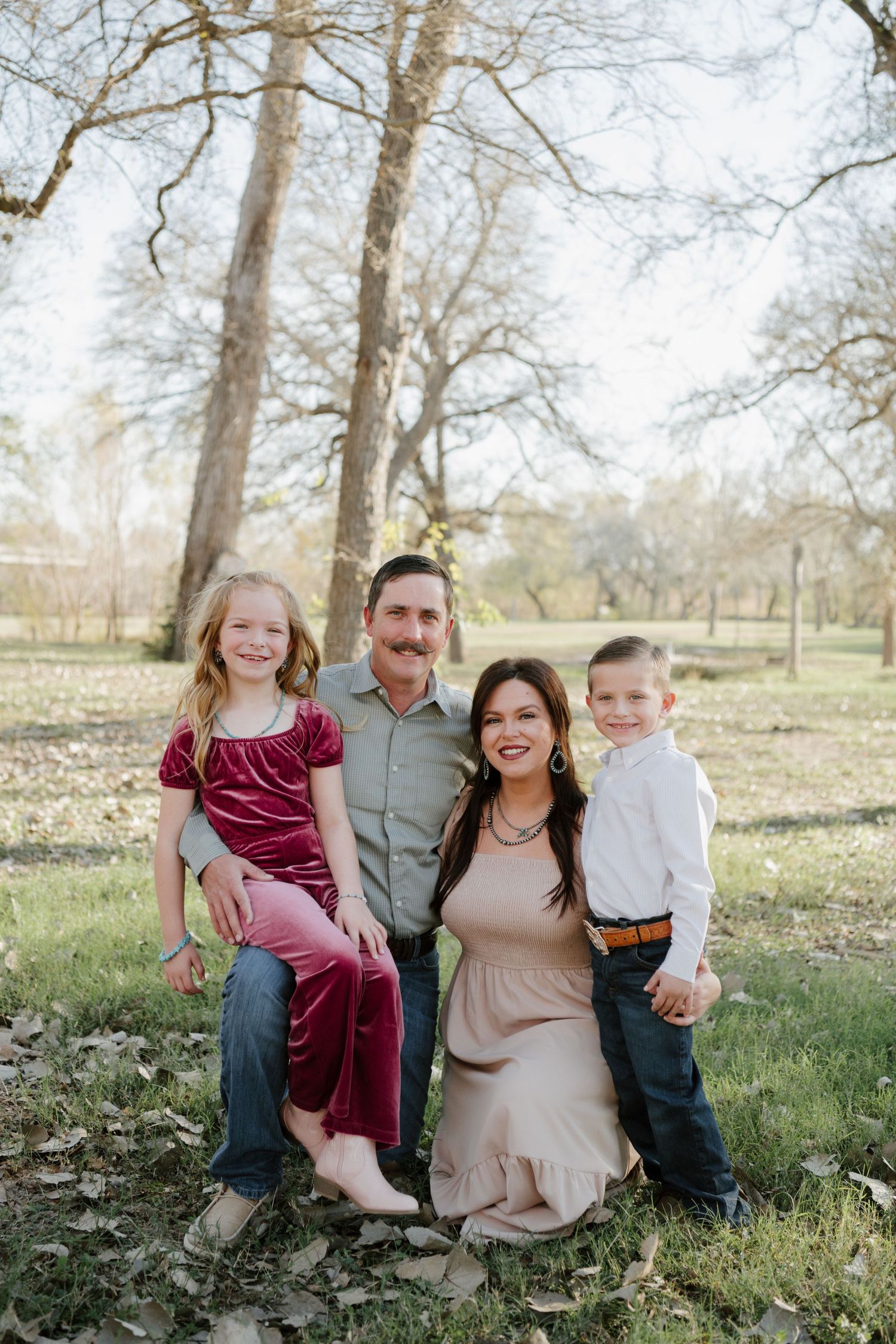 Family sitting together outdoors on grassy ground surrounded by trees in a natural park setting.