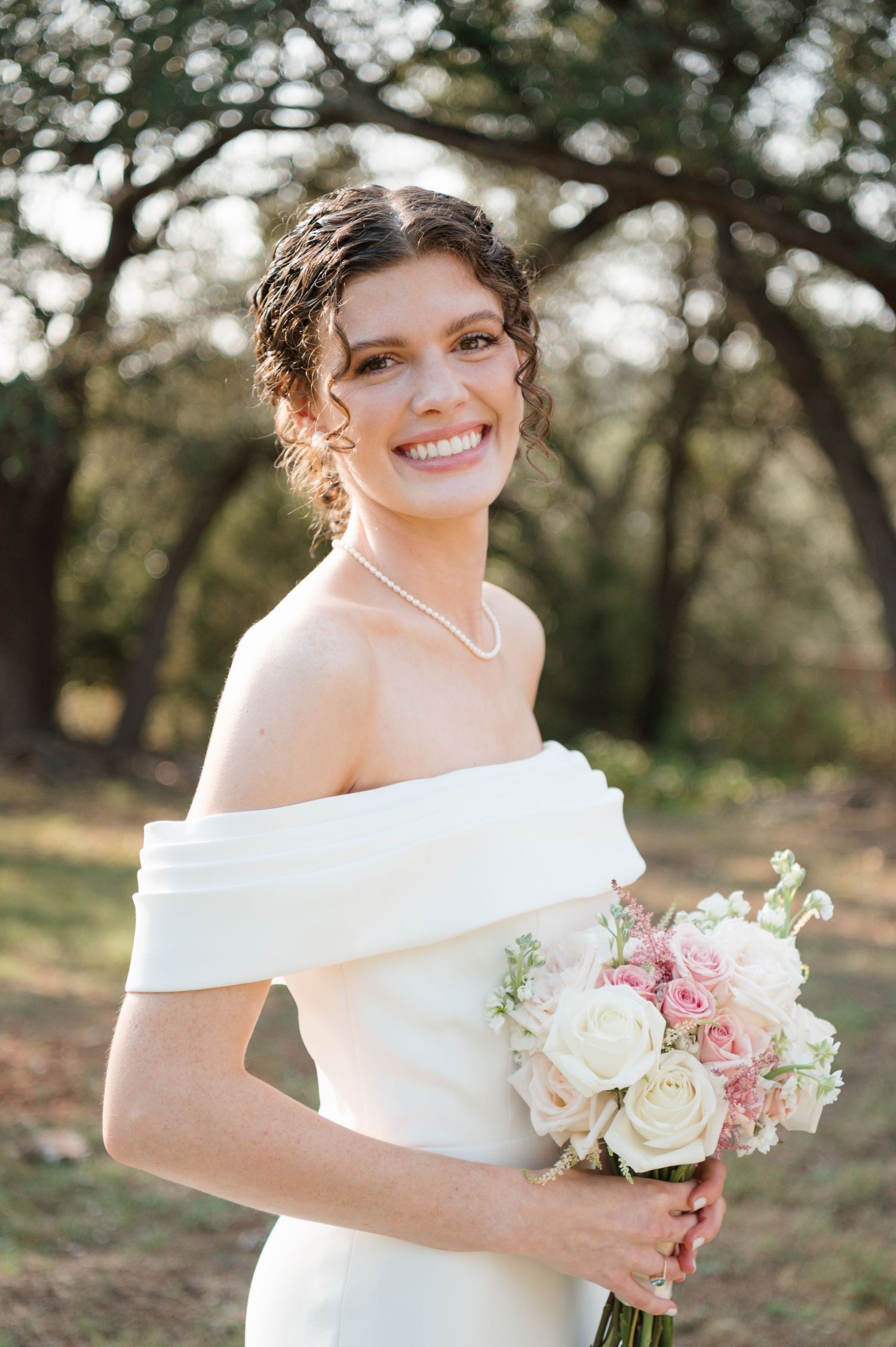 A bride in an off-shoulder white gown holds a romantic bouquet of pink and white roses in an outdoor setting.