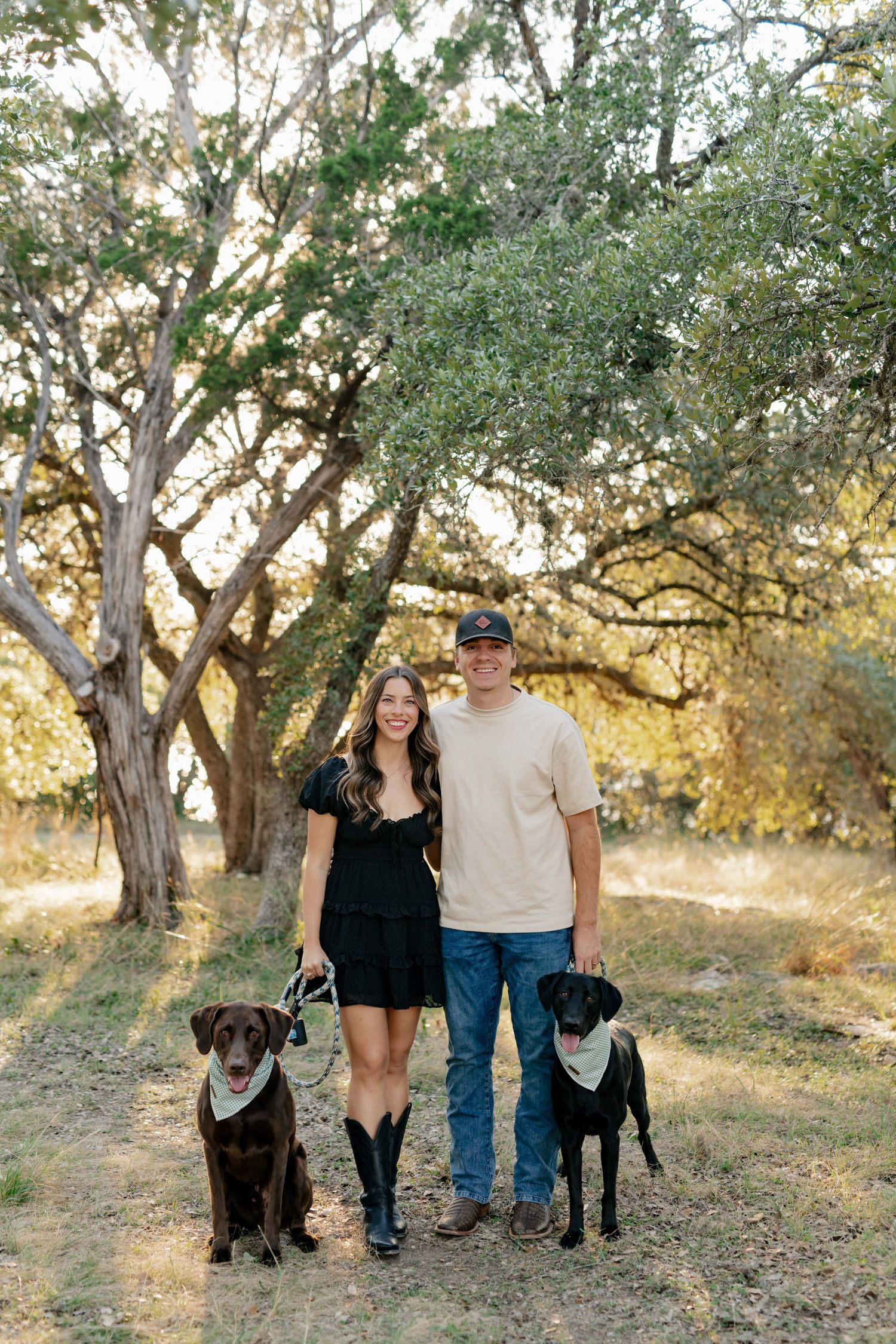 A couple poses with their two dogs on a trail surrounded by golden-lit trees in nature.